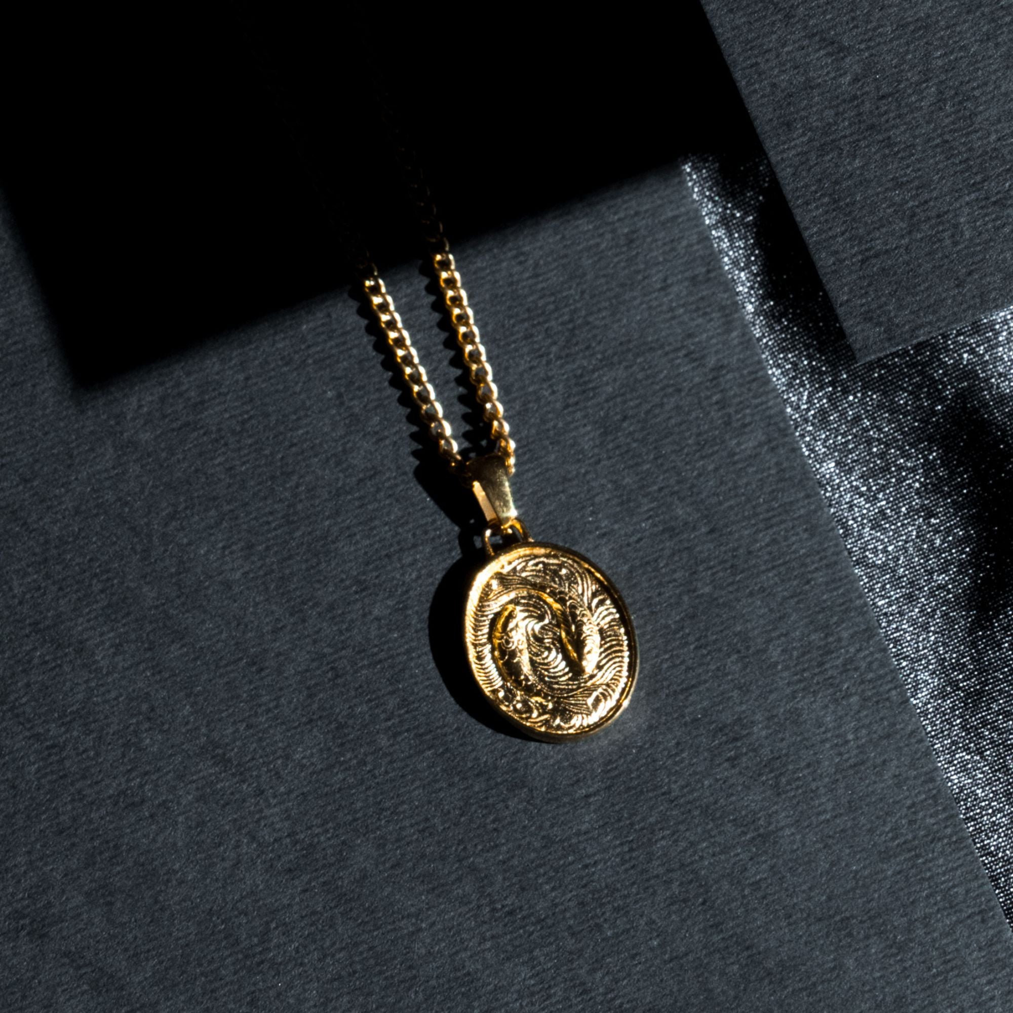 A gold coin pendant necklace with an embossed figure, resting on a dark textured surface with dramatic shadows.