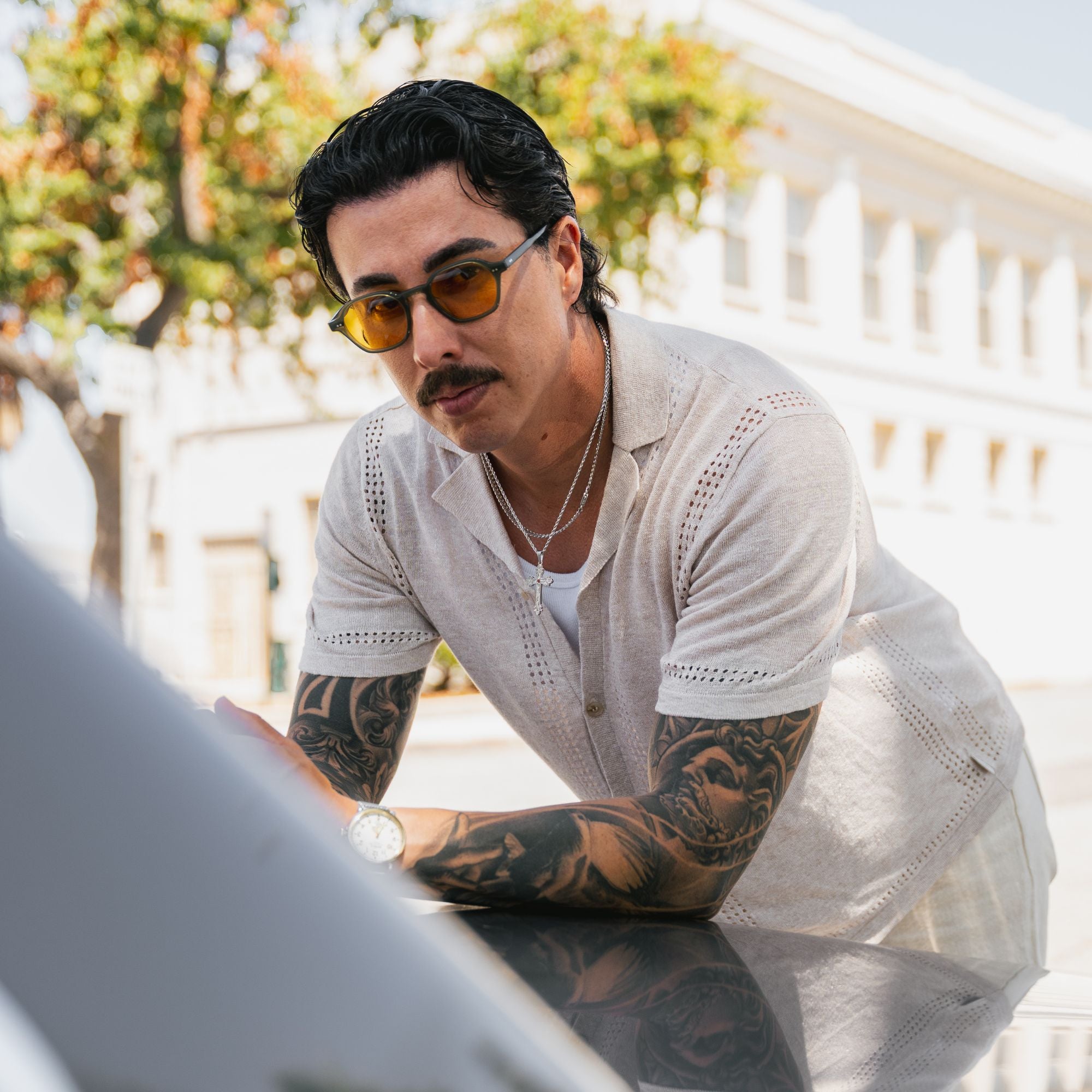 A man with a mustache and tattoos wearing yellow-tinted sunglasses, a white knit short-sleeve shirt, and a white undershirt, leaning over a reflective surface in an outdoor setting.