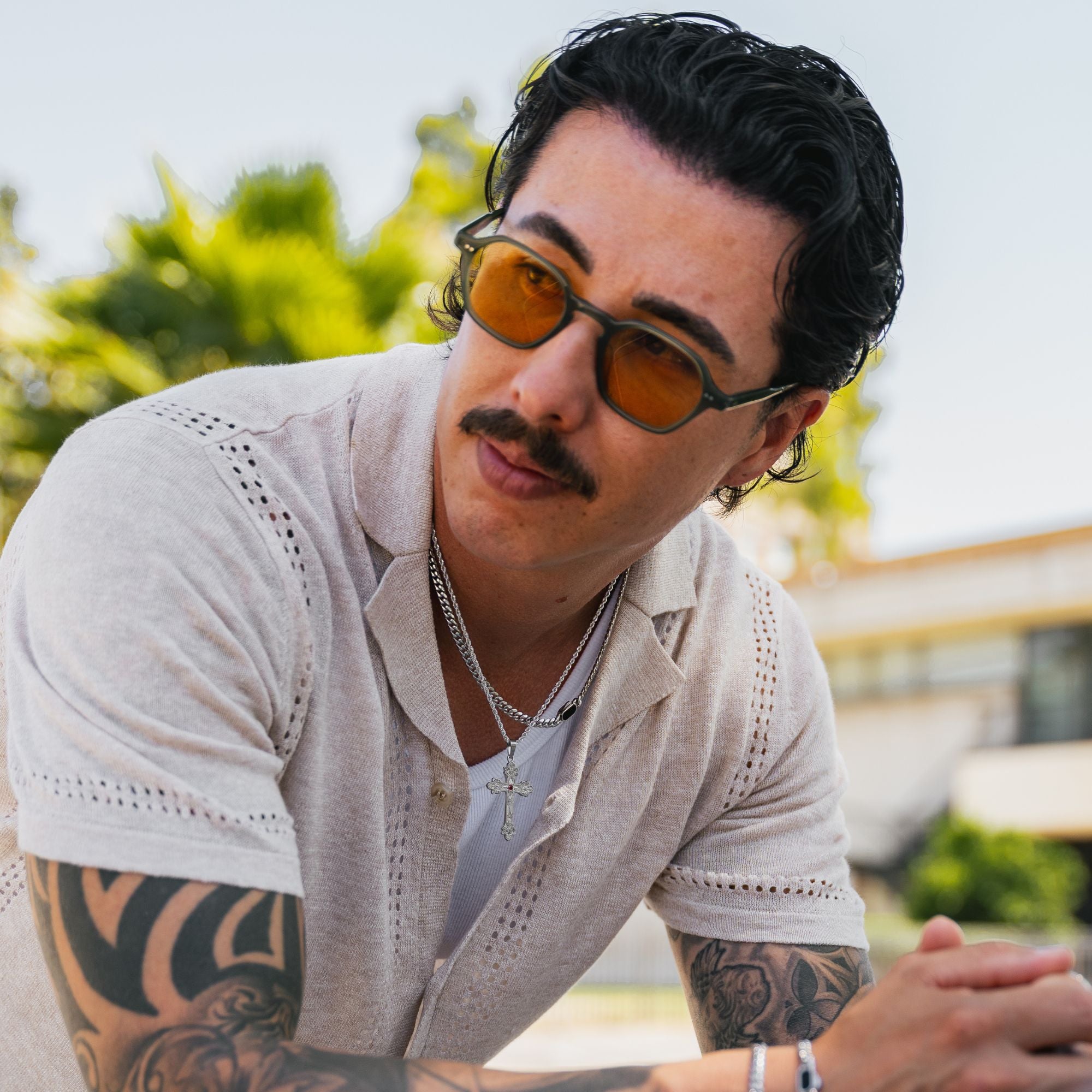 A man with dark hair and a mustache wearing orange-tinted sunglasses, a light beige short-sleeve button-up shirt, and silver necklaces, leaning forward outdoors with a blurred background of greenery and a building.