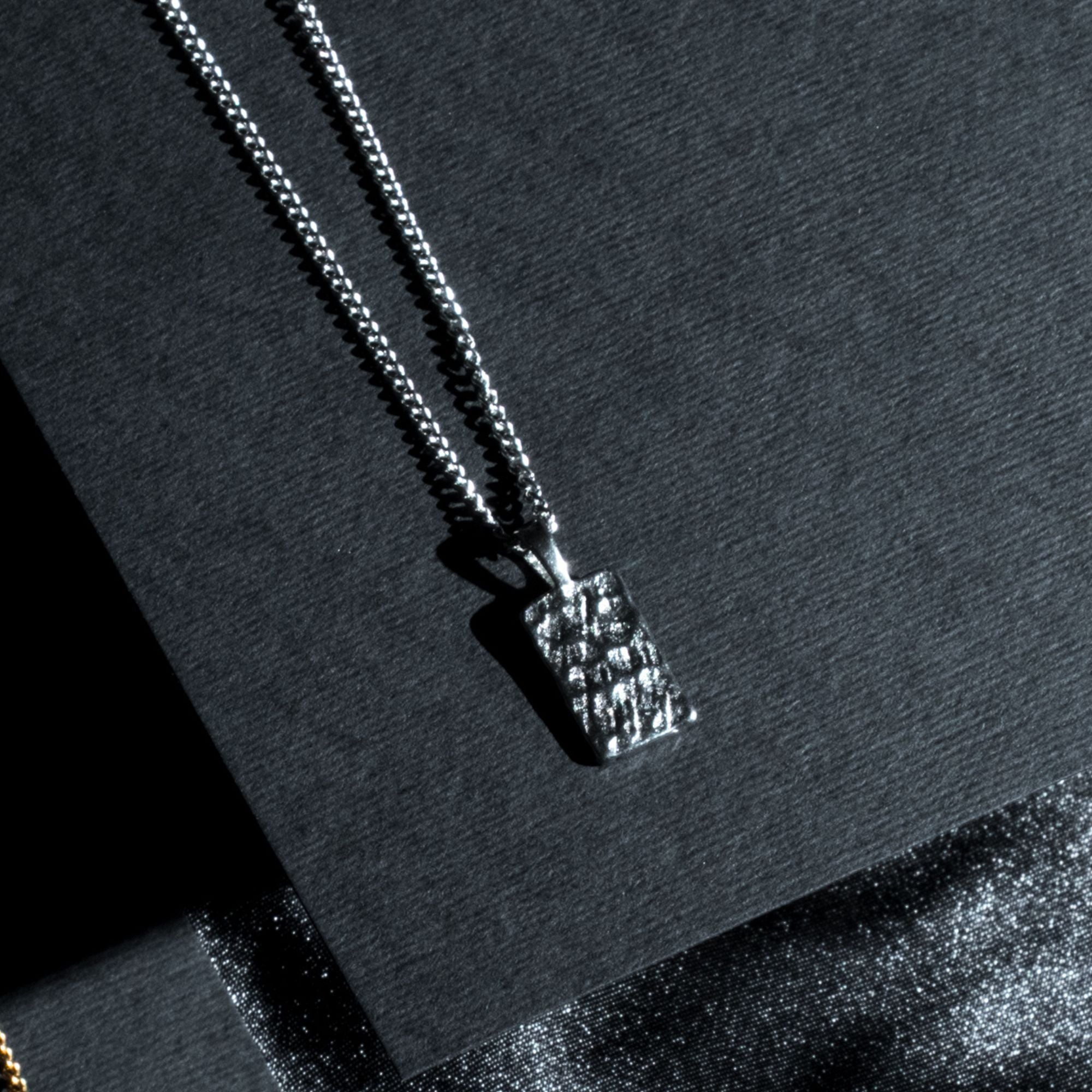 A silver-toned chain necklace with a textured rectangular pendant, displayed on a dark charcoal background.