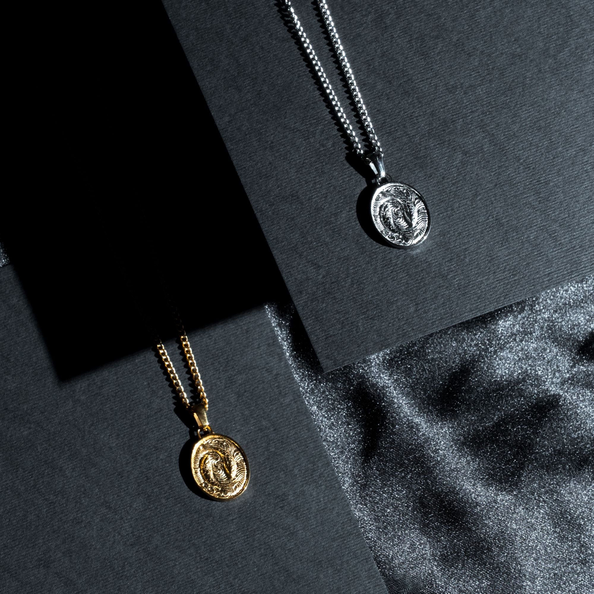Two coin pendant necklaces displayed on dark textured cards, one in gold and one in silver, with a dark gray fabric surface in the background.