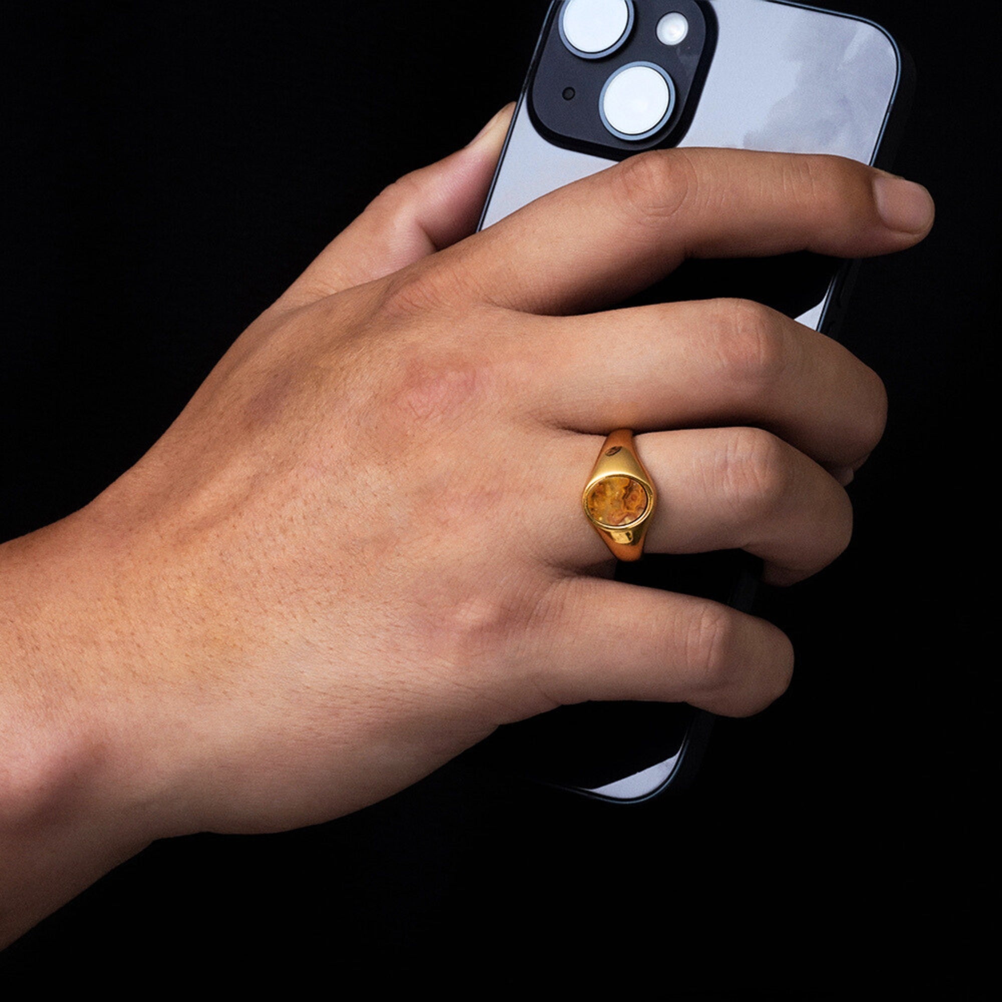 Hand wearing a gold ring holding a smartphone on a black background