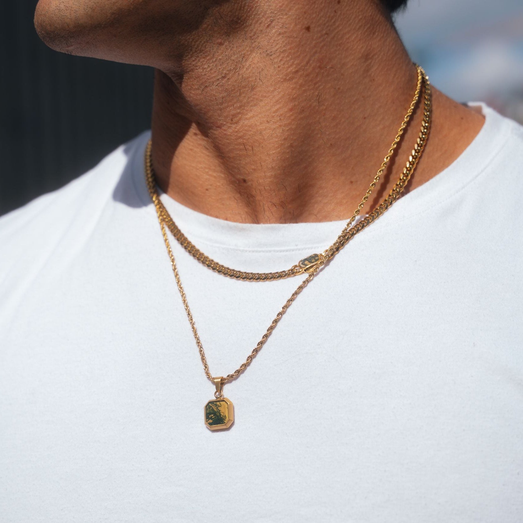 A close-up of a person's neck and chest wearing a layered gold necklace set, featuring a curb chain and a delicate chain with a small square pendant, over a white t-shirt.