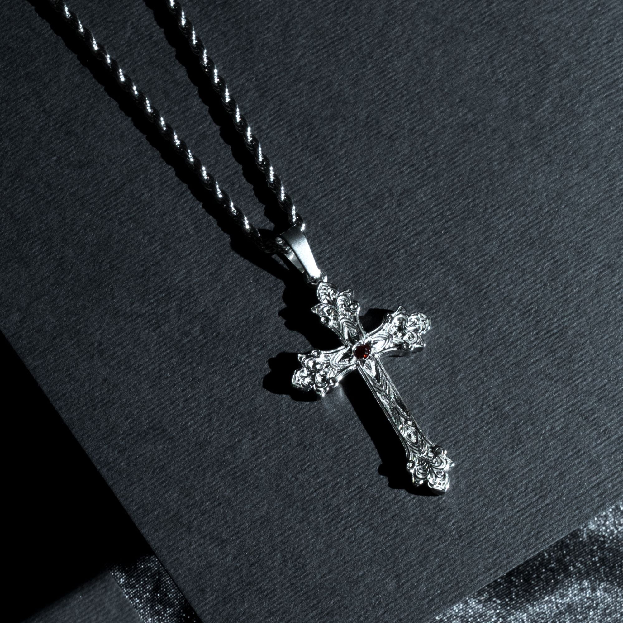 A silver-toned ornate cross pendant necklace with a small red gemstone at the center, resting on a textured black surface.