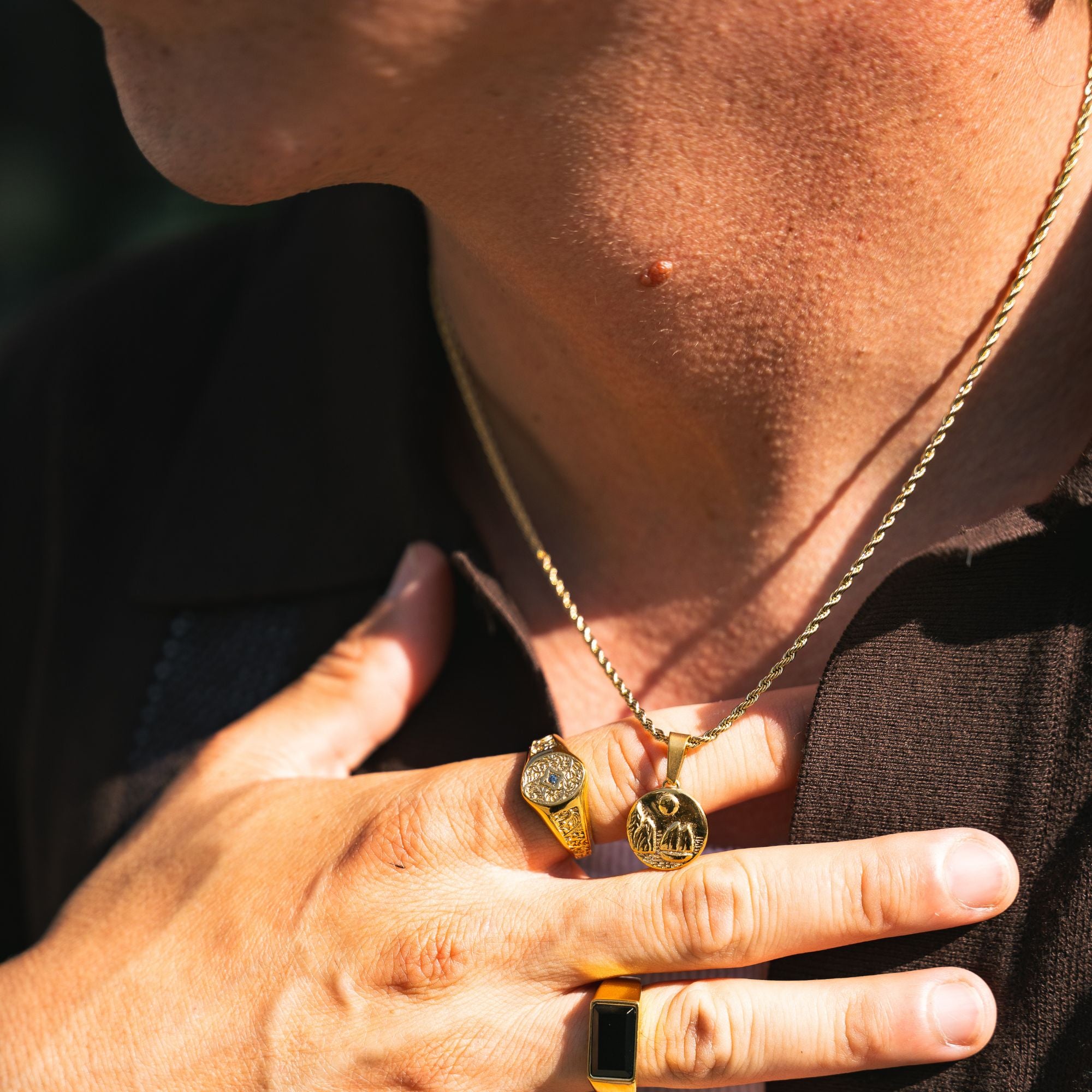 A close-up lifestyle shot of a person wearing a gold chain necklace with two small gold pendants, one of which features a circular emblem. The person is also wearing gold rings, including a prominent black and gold signet-style ring on the index finger.