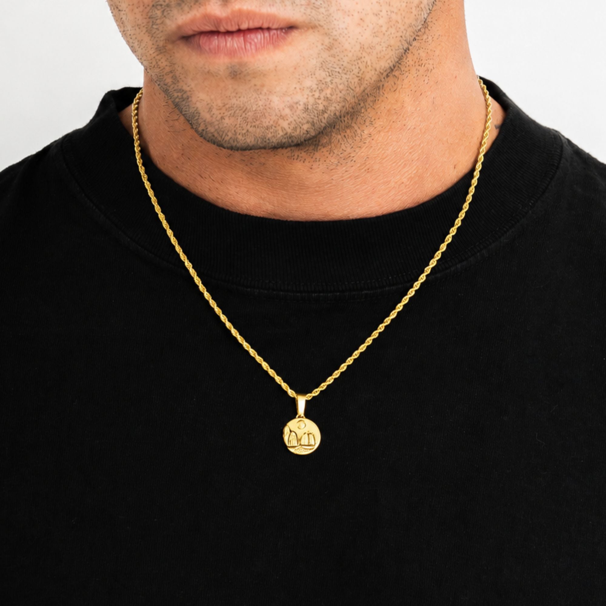 A close-up of a man wearing a gold chain necklace with a round engraved gold pendant, styled with a black crewneck sweatshirt against a light gray background.