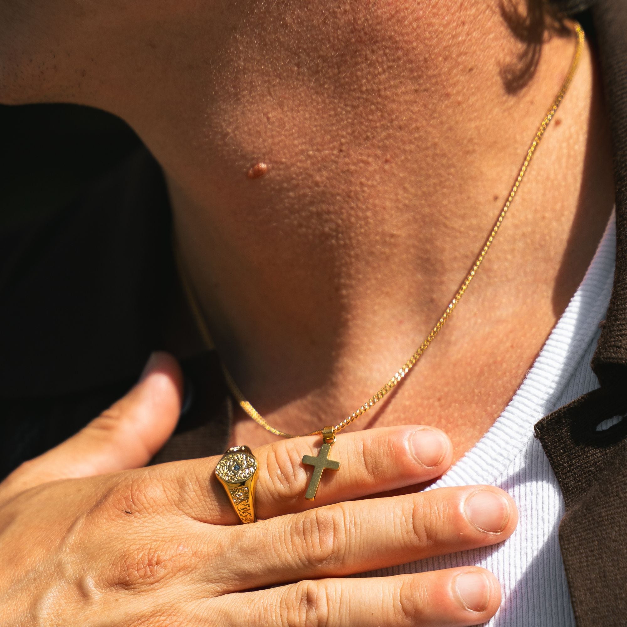 A close-up lifestyle shot of a person's neck and hand, showcasing a delicate gold chain necklace with a small cross pendant resting on their chest, alongside a gold ring on their finger.