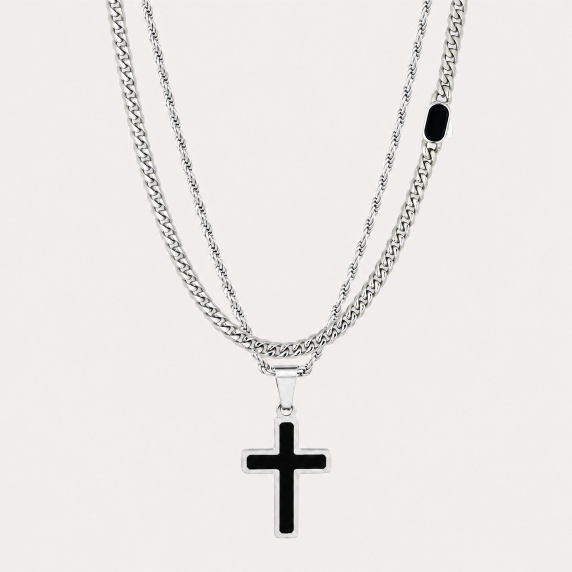 A double-layered necklace featuring a silver-toned chain with a black cross pendant and a textured silver chain with a small black oval accent, set against a light off-white background.