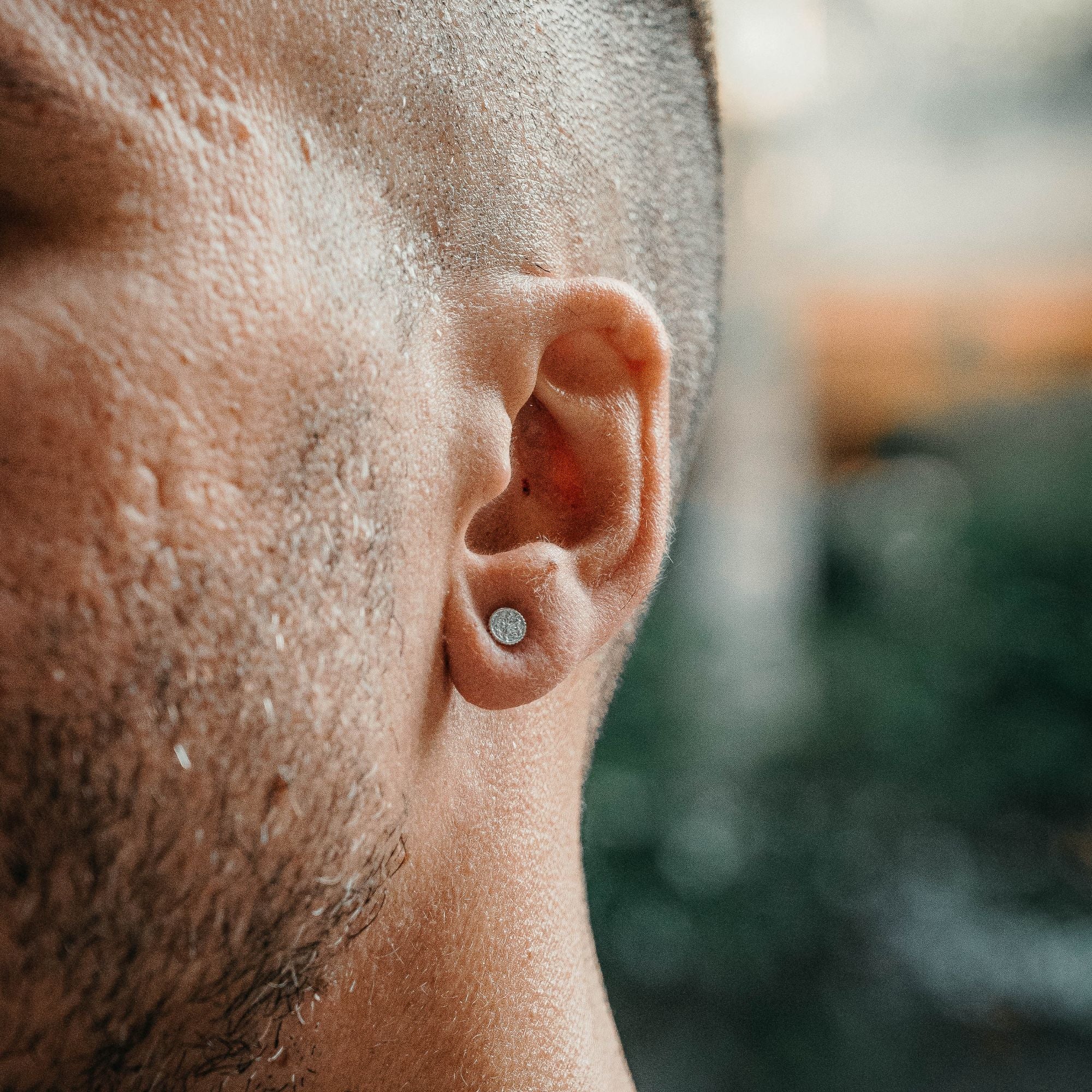A close-up side profile of a man's ear wearing a small silver stud earring, with his skin and short hair filling the foreground against a blurred outdoor background.