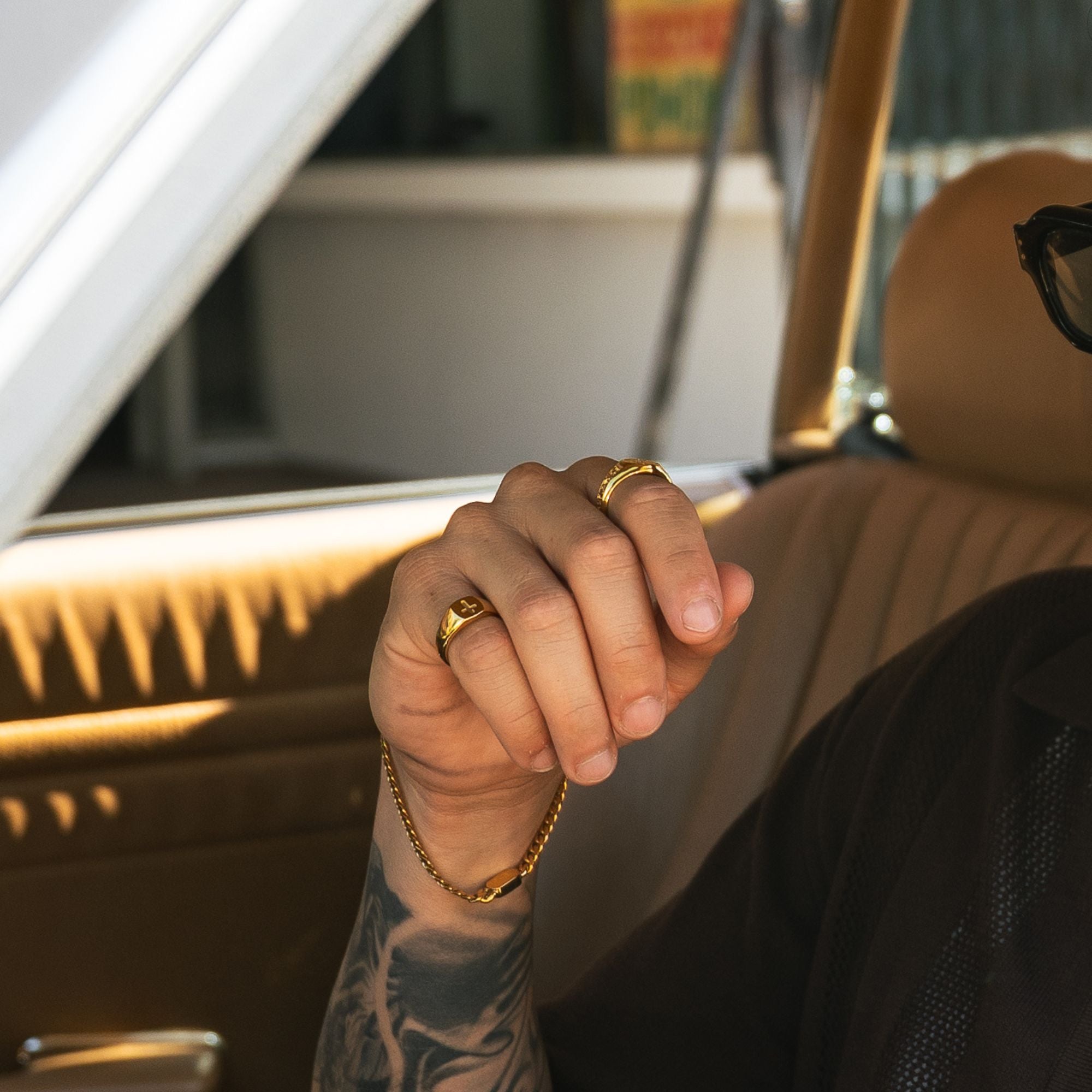A close-up of a person's hand resting inside a car, wearing two gold rings and a gold chain bracelet. The person is wearing a black shirt and sunglasses, with a tan leather car interior in the background.