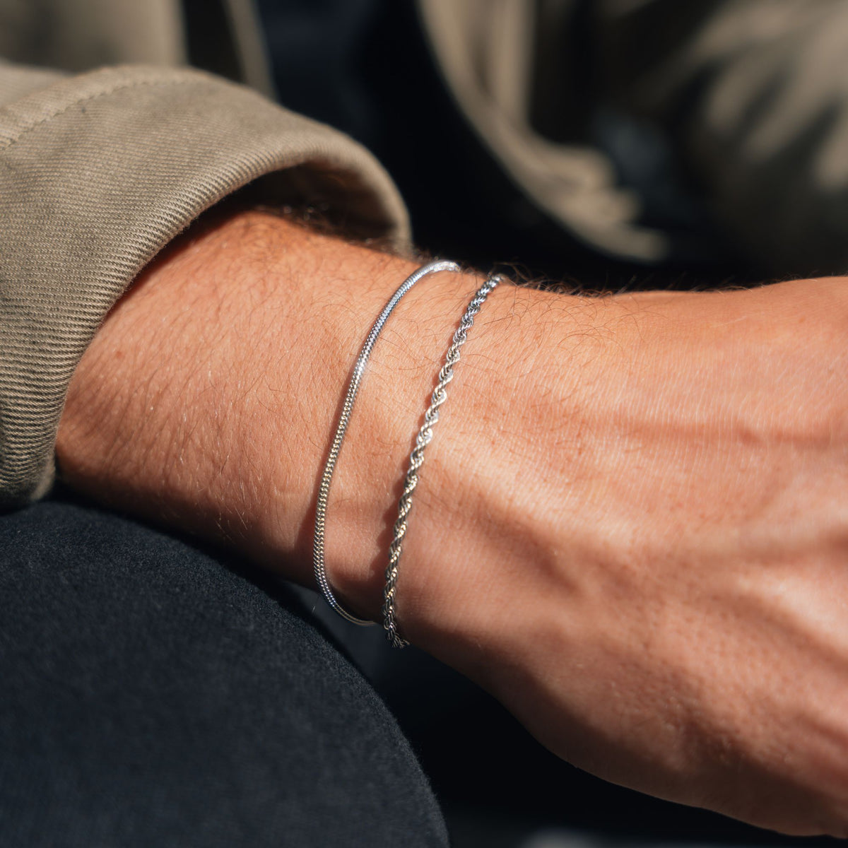 Two silver bracelets, one thin snake chain and one twisted rope chain, worn on a person's wrist.