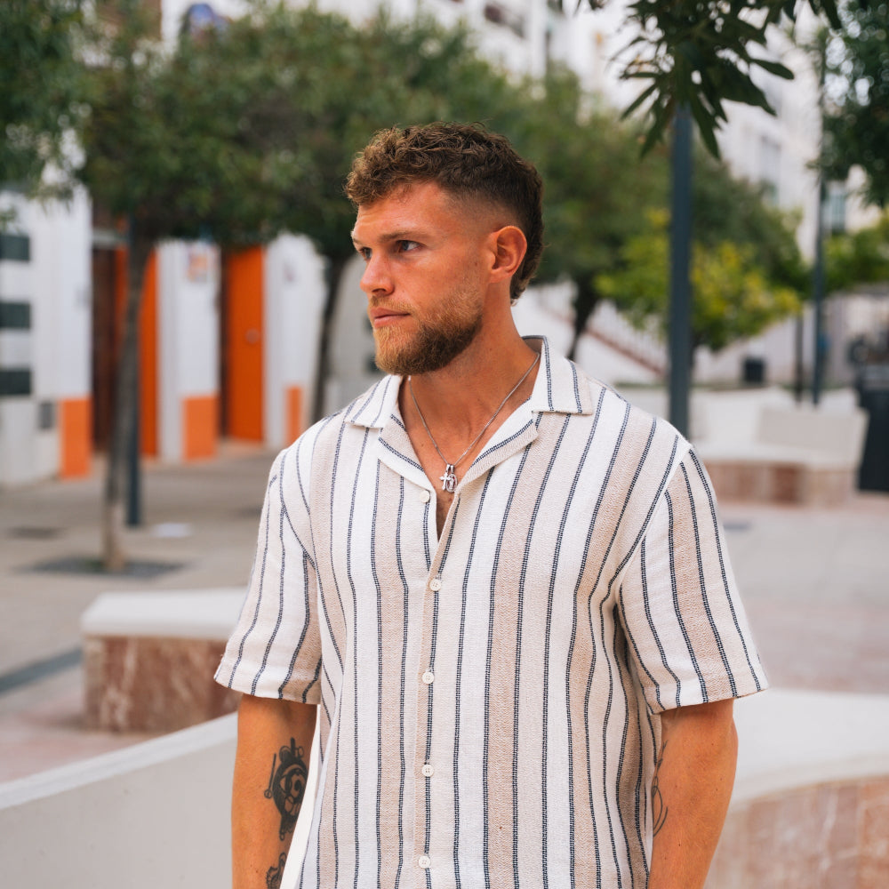 Man wearing a striped shirt standing outdoors with trees and buildings in the background