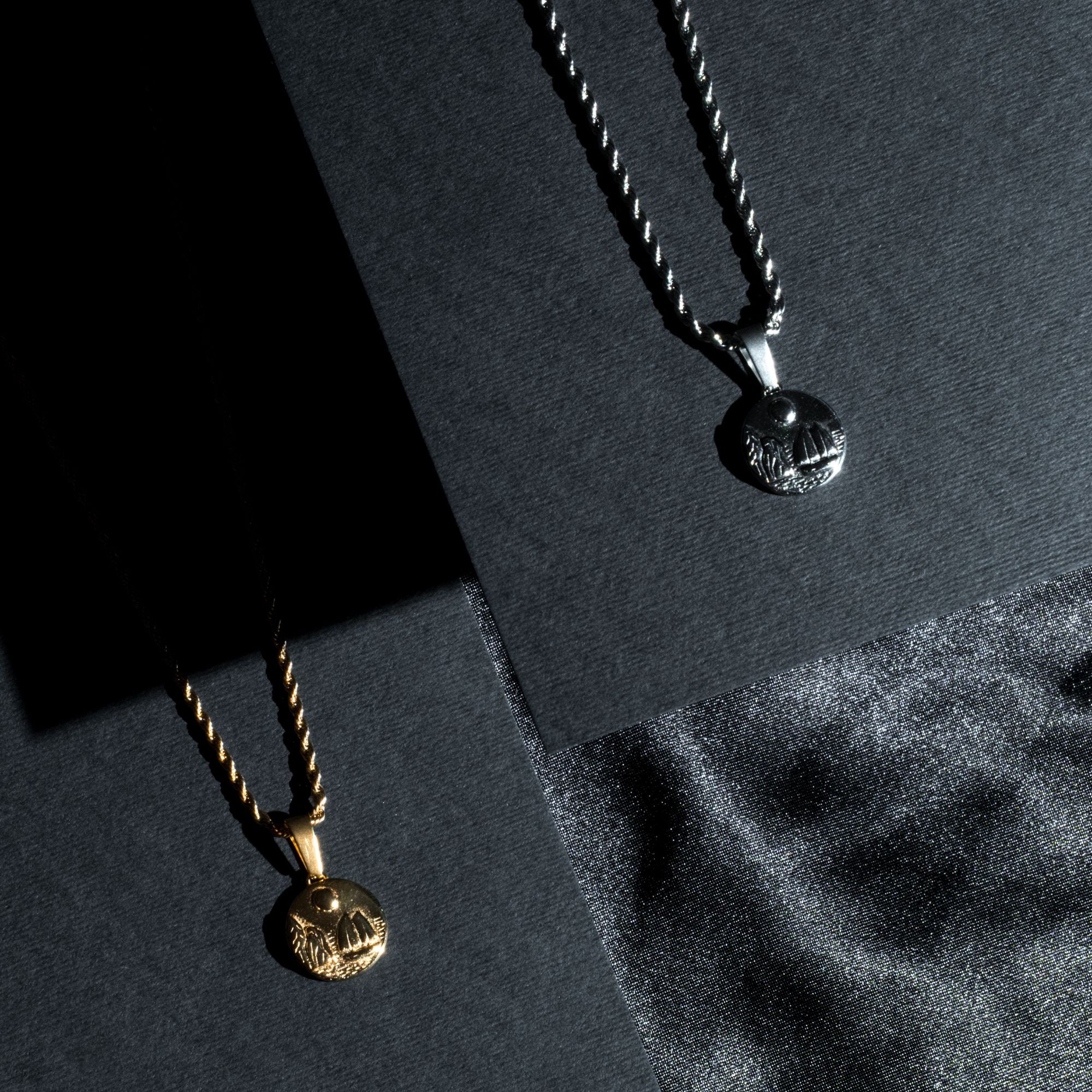 Two pendant necklaces displayed on dark textured panels: one gold-toned with a round engraved pendant and twisted chain, and one silver-toned with a matching round pendant and twisted chain.