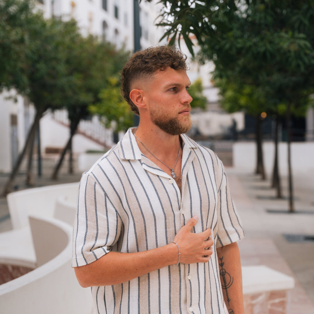 Man wearing a striped shirt standing outdoors with trees and buildings in the background