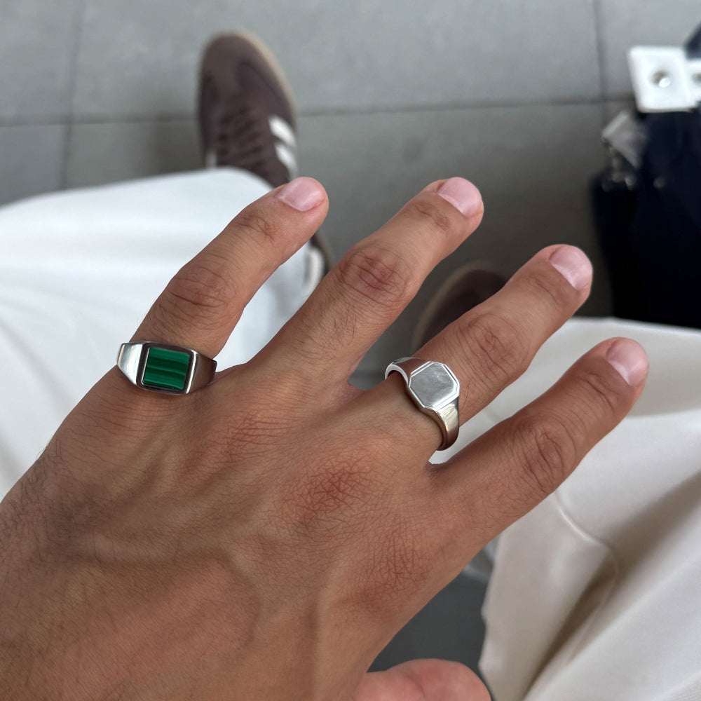 Hand wearing two rings with a green gemstone and silver band on a neutral background