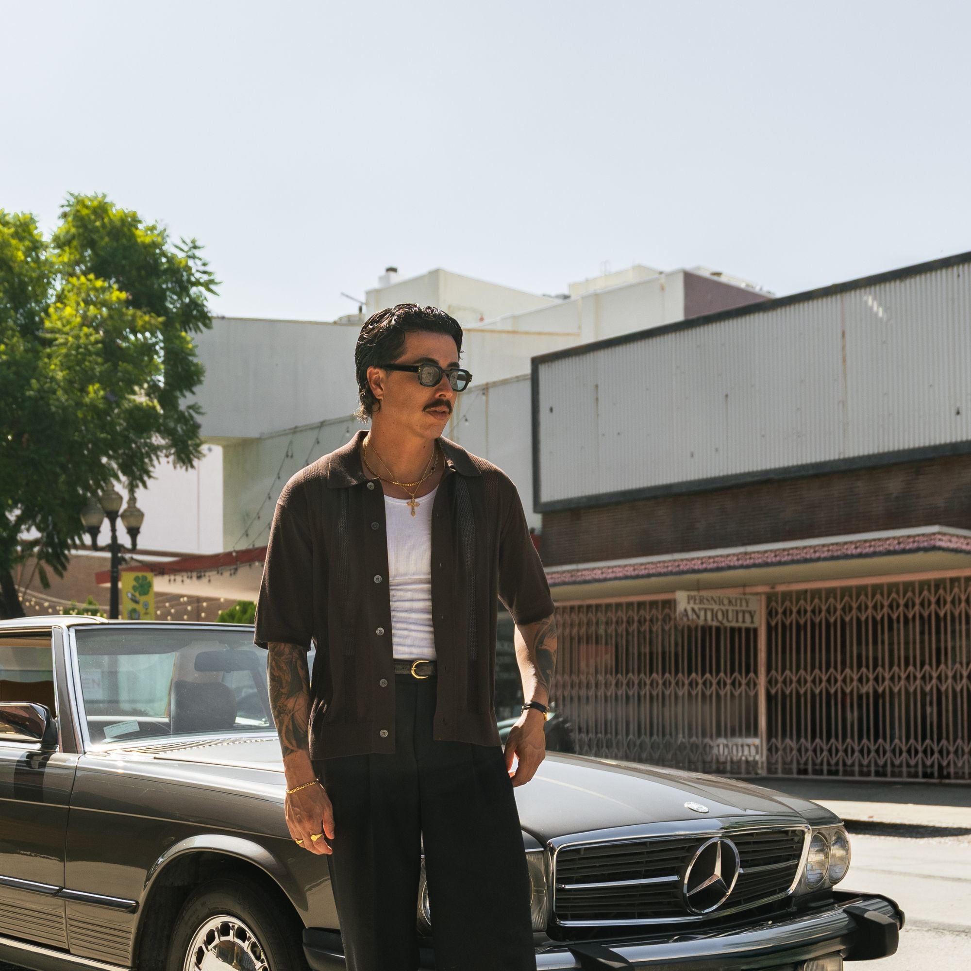 A man wearing a dark brown short-sleeve button-up shirt, black pants, and sunglasses stands next to a black Mercedes-Benz car on a sunny street.