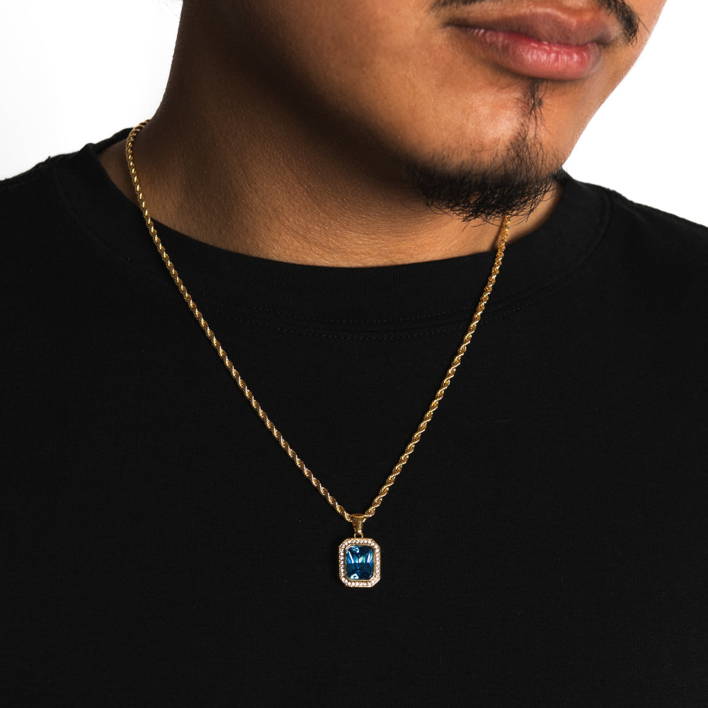 Man wearing a gold necklace with a blue gemstone pendant on a white background