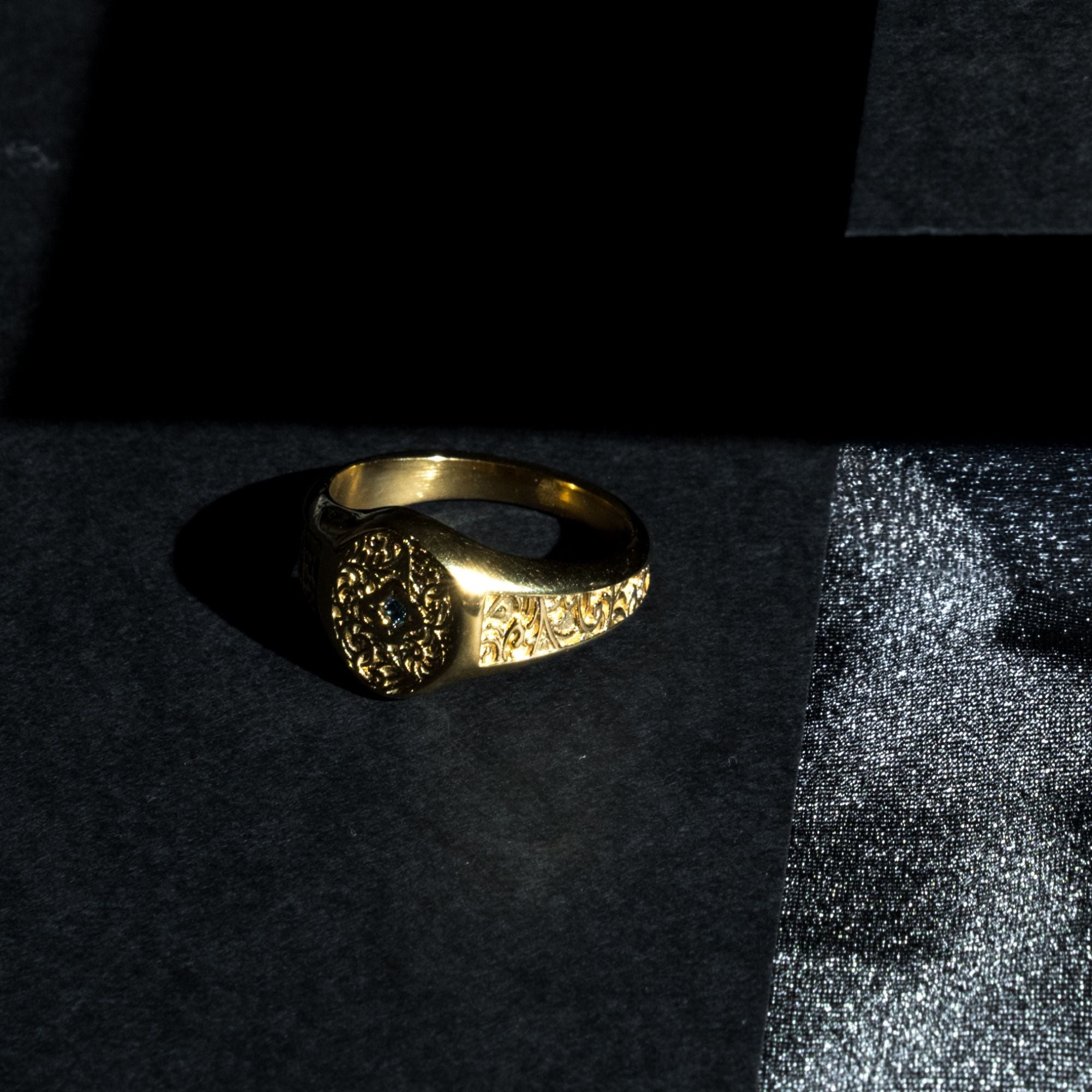 A textured gold signet ring with an ornate floral or scrollwork face, resting on a dark textured surface with dramatic side lighting.