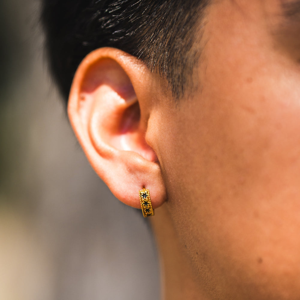 Close-up of an ear wearing a gold hoop earring with black stones against a blurred background