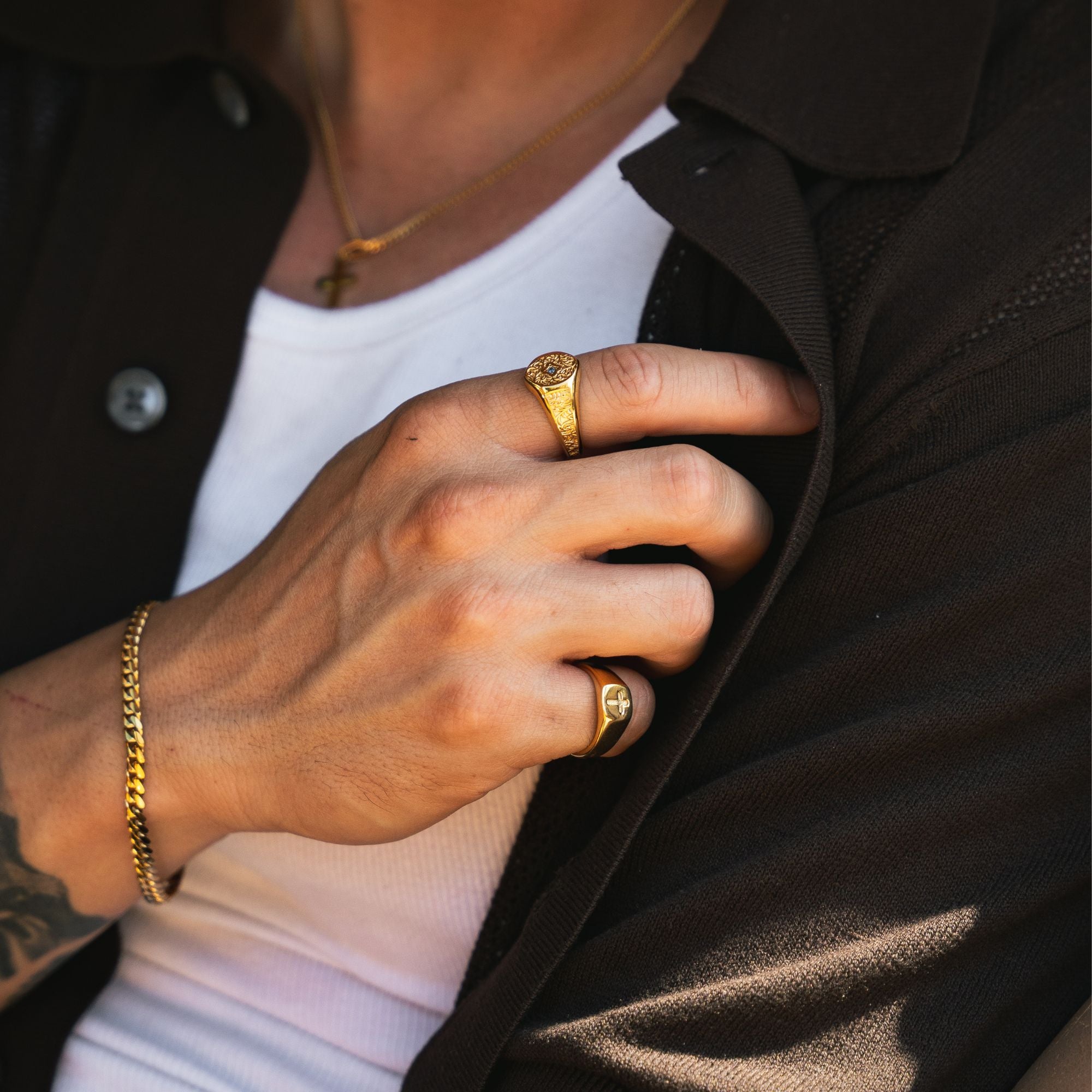 A close-up lifestyle shot of a person wearing gold jewelry, featuring a gold signet ring on one finger, a gold chain bracelet on the other, and a gold necklace. The person is wearing a white t-shirt and a dark brown collared shirt.