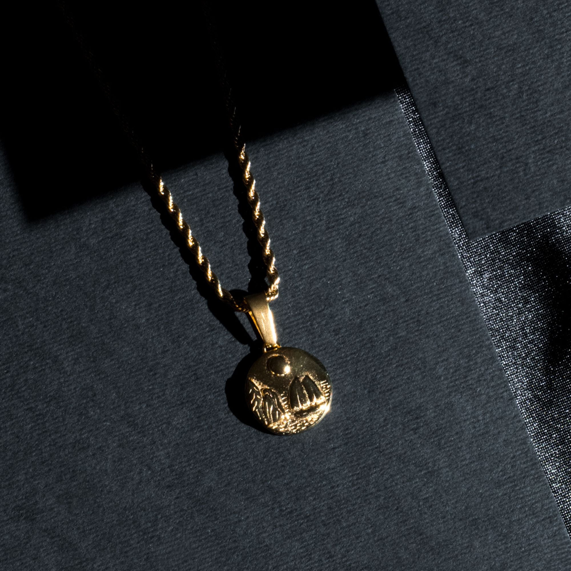 A gold pendant necklace with a textured circular medallion pendant, resting on a dark gray textured surface with dramatic shadow lighting.