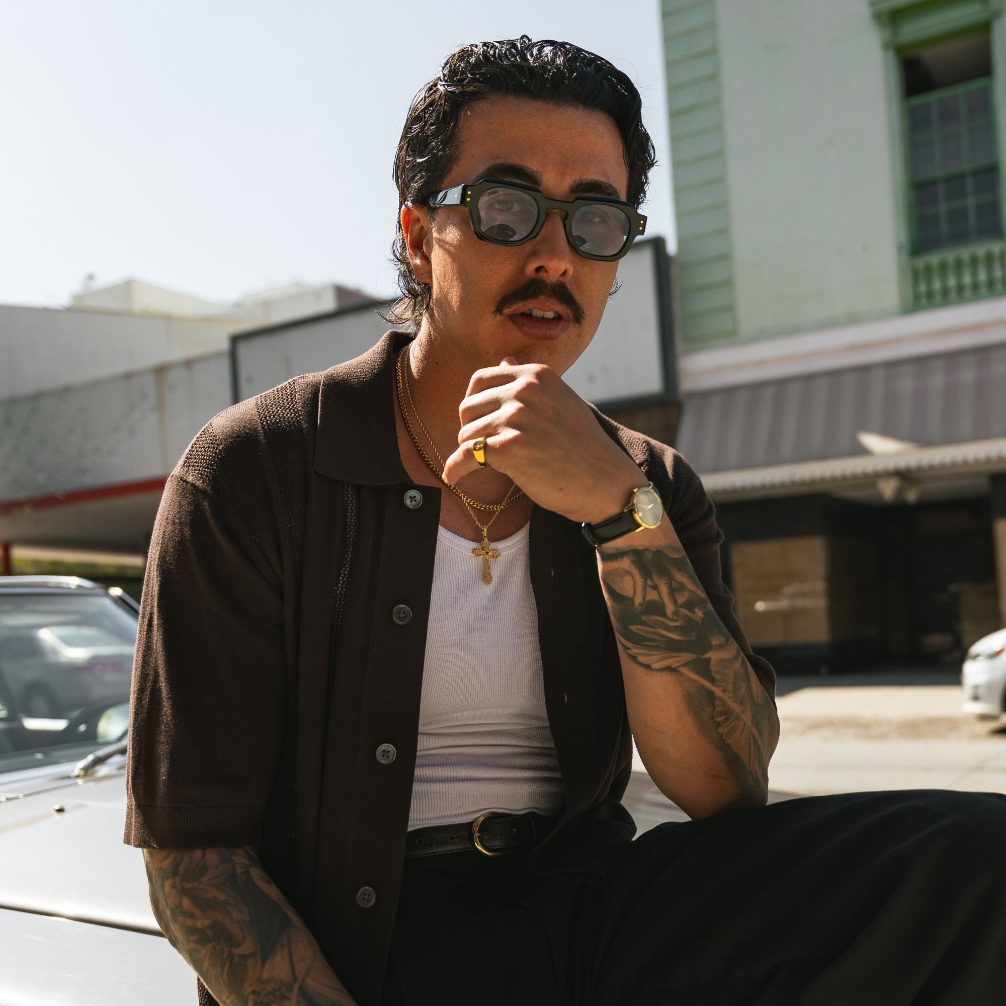 A man with tattoos and a mustache wearing dark sunglasses, a dark brown short-sleeve button-up shirt over a white tank top, and black pants, sitting outdoors in front of a building.