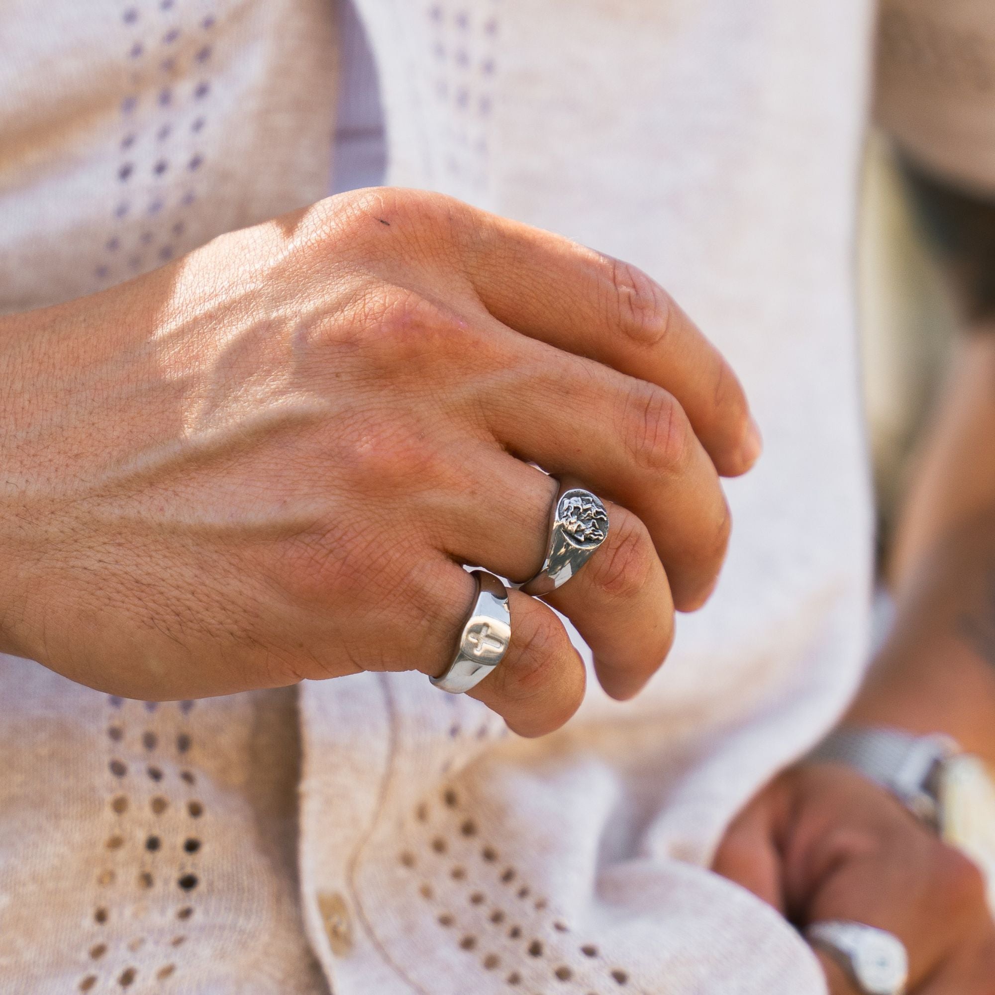 A close-up of a person's hand wearing two silver rings, one a simple band and the other a signet-style ring with an engraved floral or scroll design, set against a light cream knit garment.