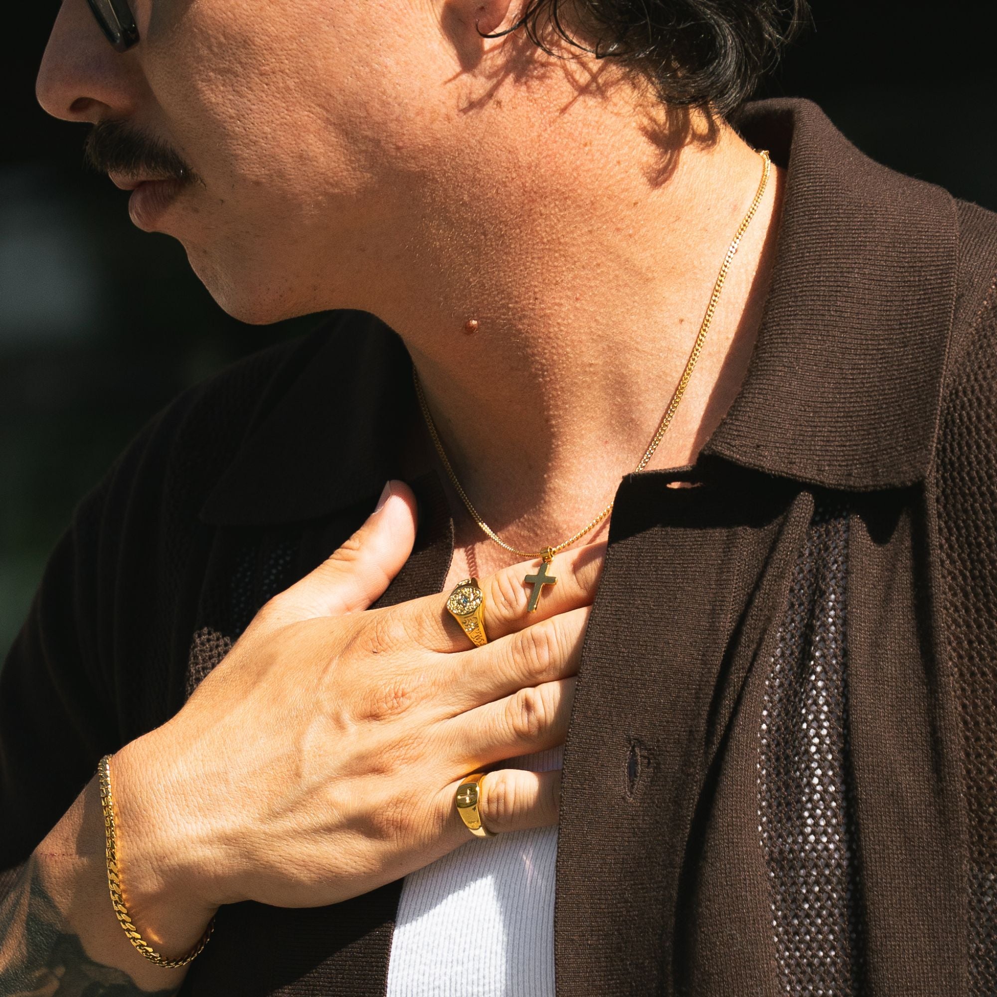 Close-up of a man wearing gold jewelry, including a gold chain necklace, a gold ring, and a gold bracelet, with his hand resting on his chest.