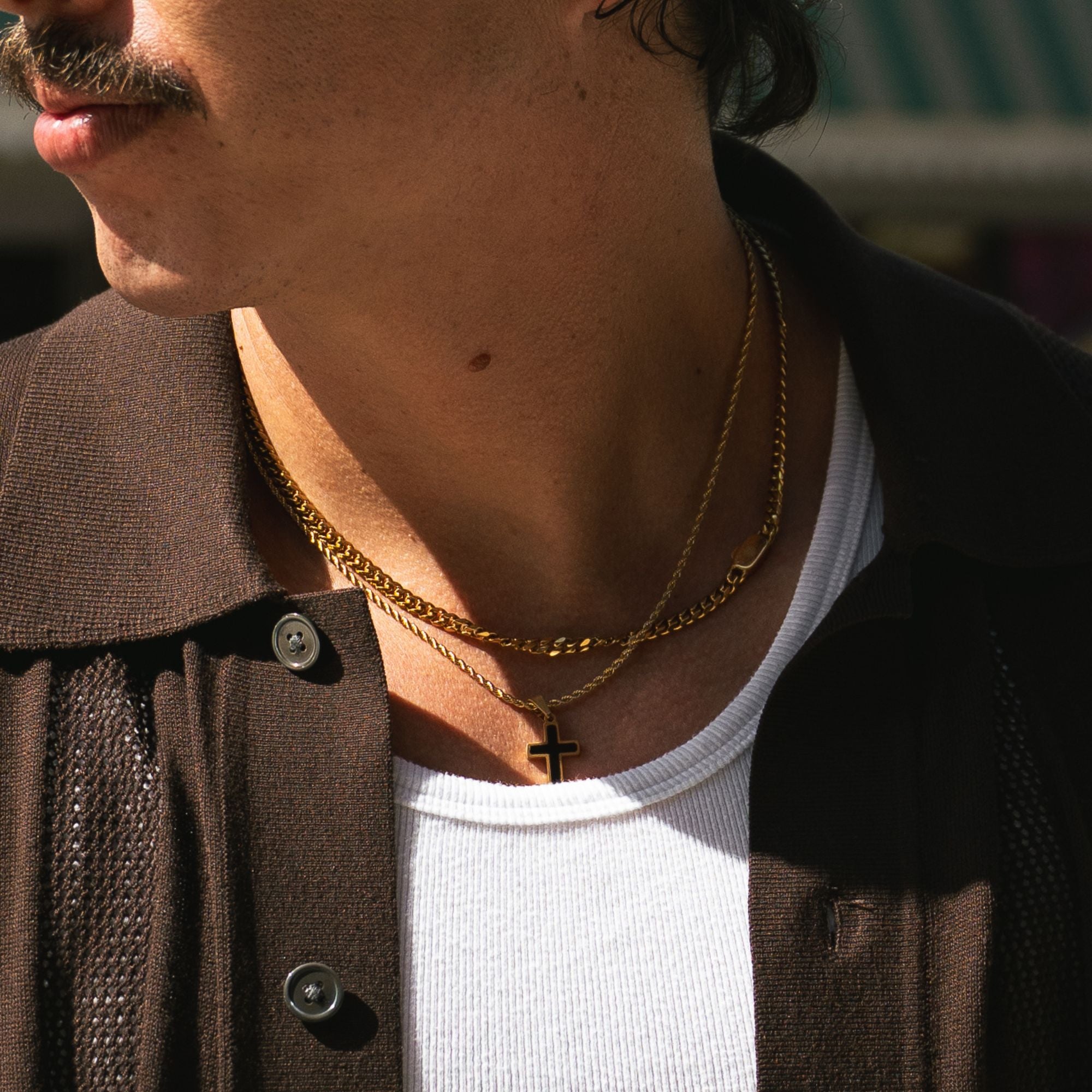 A close-up lifestyle shot of a person wearing layered gold necklaces, including a curb chain and a delicate chain with a small cross pendant, over a white t-shirt and dark brown jacket.