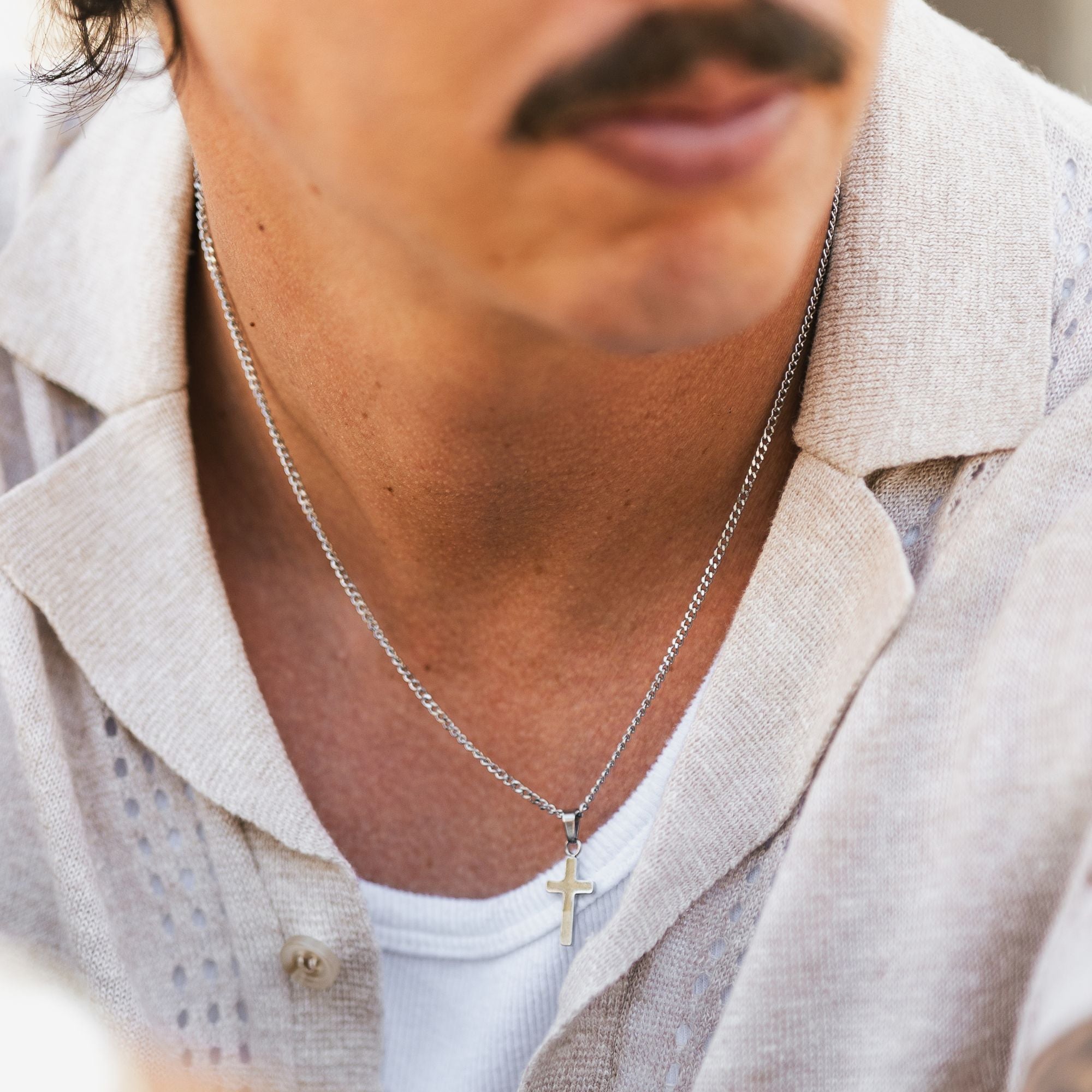 A close-up of a man wearing a silver chain necklace with a small cross pendant, styled with a light beige linen shirt over a white undershirt.