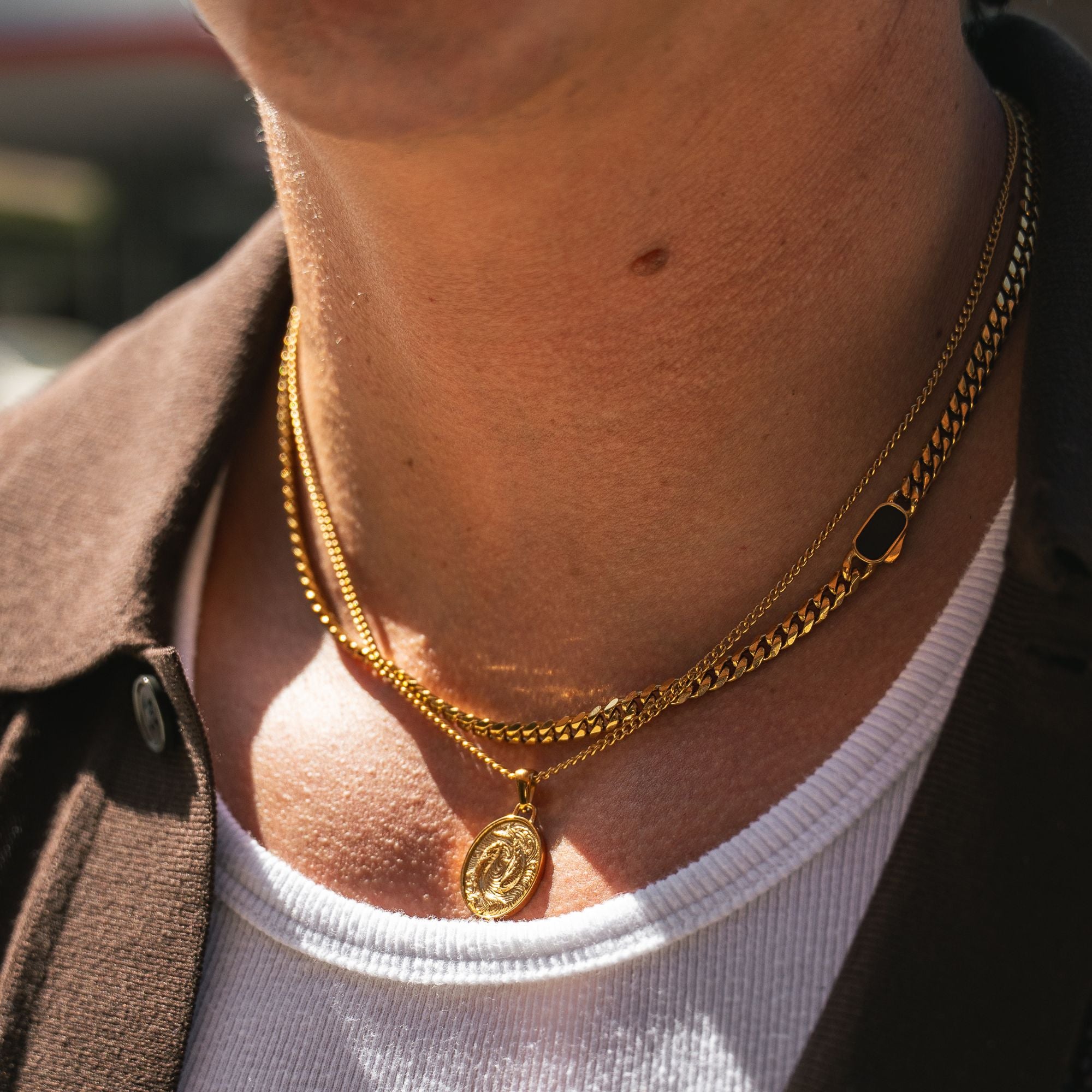 A close-up of a person's neck wearing two layered gold necklaces: a thin chain with a round coin-style pendant and a slightly thicker chain with a small dark rectangular accent.