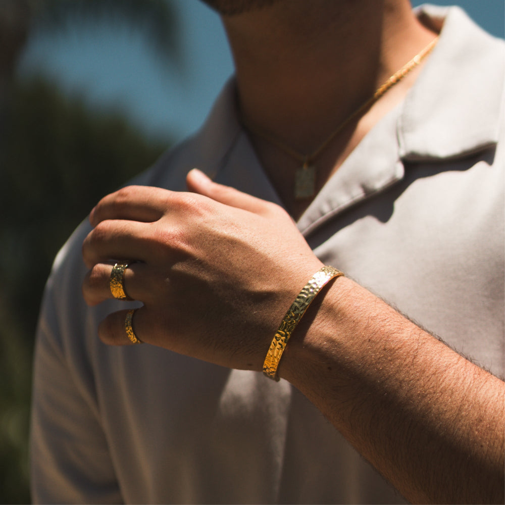 Close-up of a person wearing gold jewelry including a ring and bracelet, with a blurred natural background.