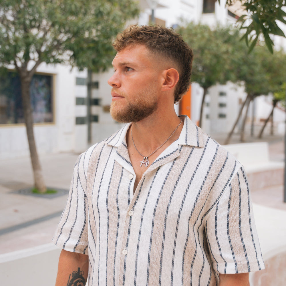Man wearing a striped shirt standing outdoors with trees and buildings in the background