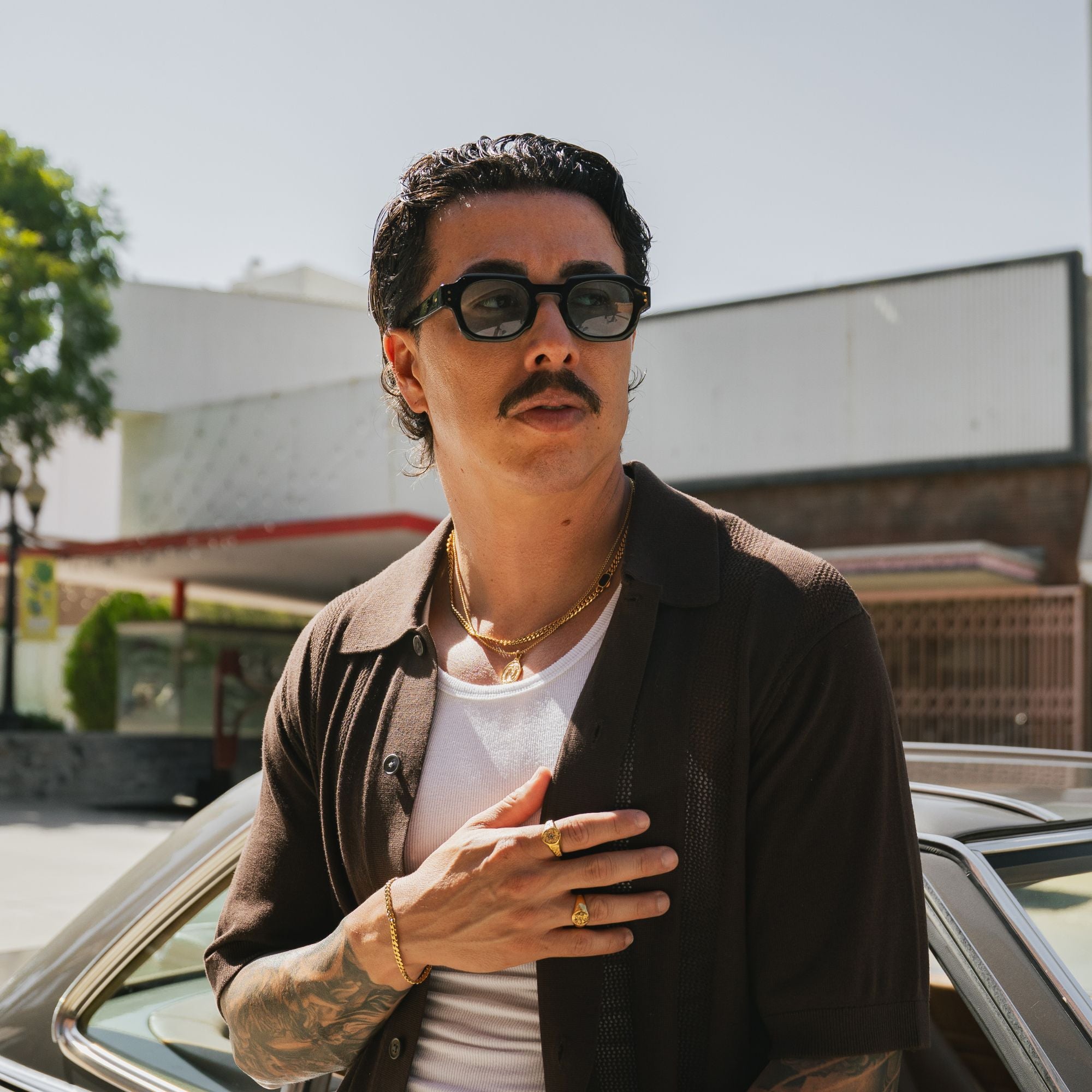 A man wearing black sunglasses, a dark brown button-up shirt over a white t-shirt, and gold jewelry, standing outdoors next to a car.