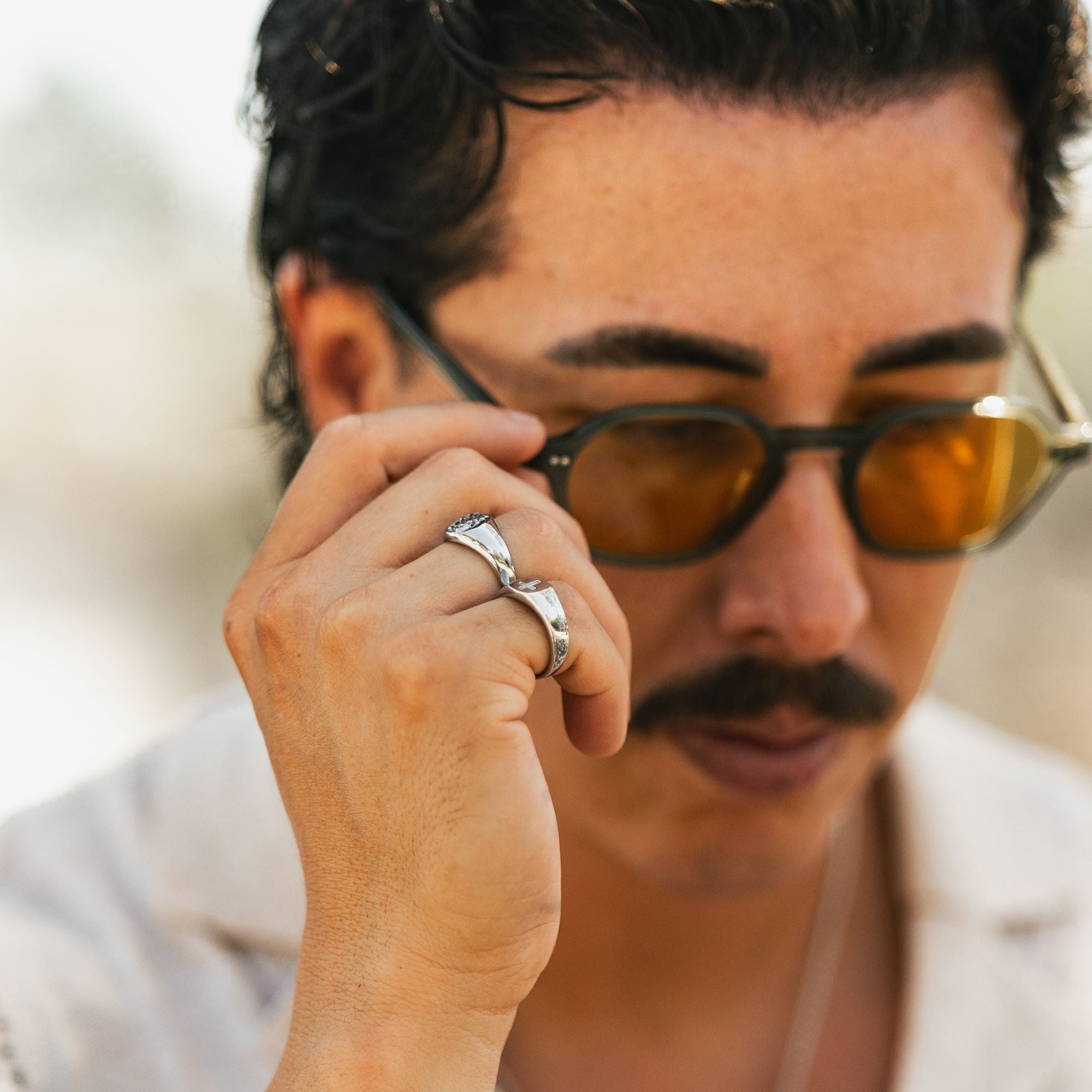 A close-up lifestyle shot of a man wearing black sunglasses with amber-tinted lenses and silver rings on his fingers, holding one lens with his hand.