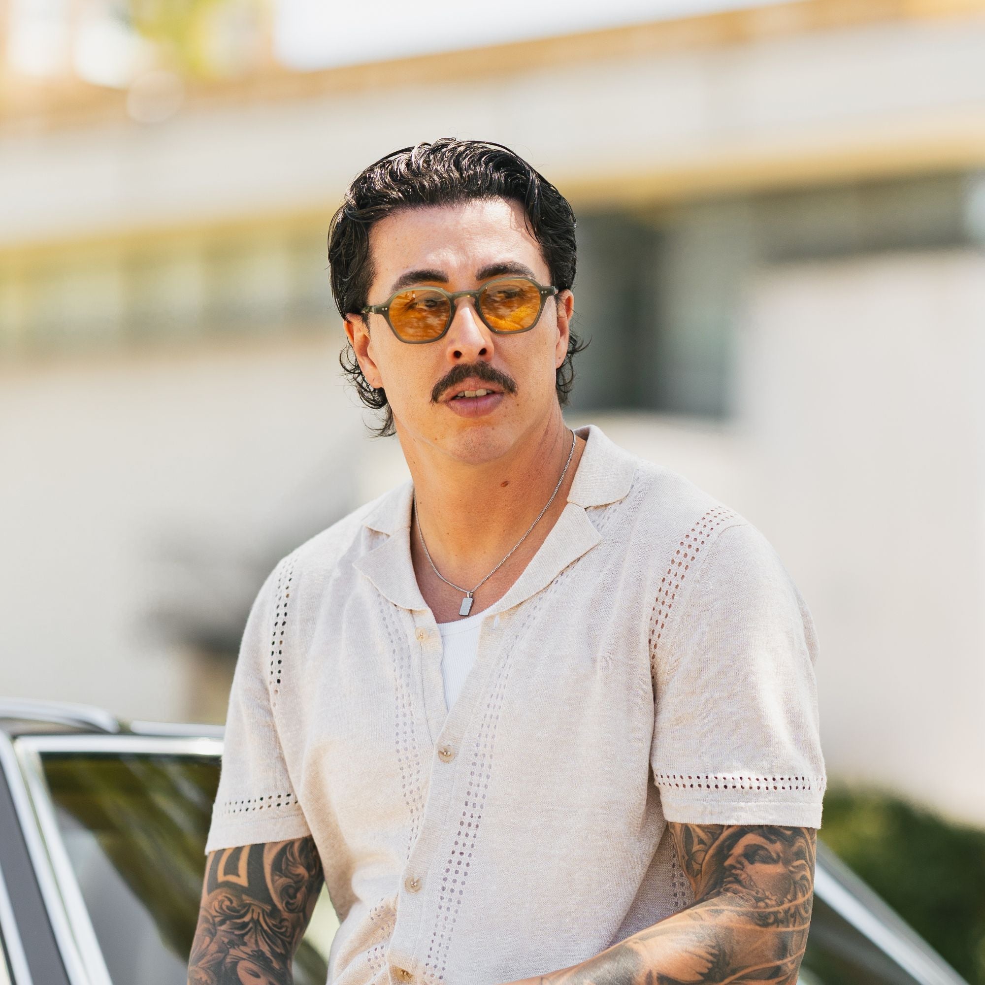A man with a mustache and tattoos wearing yellow-tinted sunglasses, a white t-shirt, and a beige short-sleeve knit button-up shirt, standing outdoors near a car.