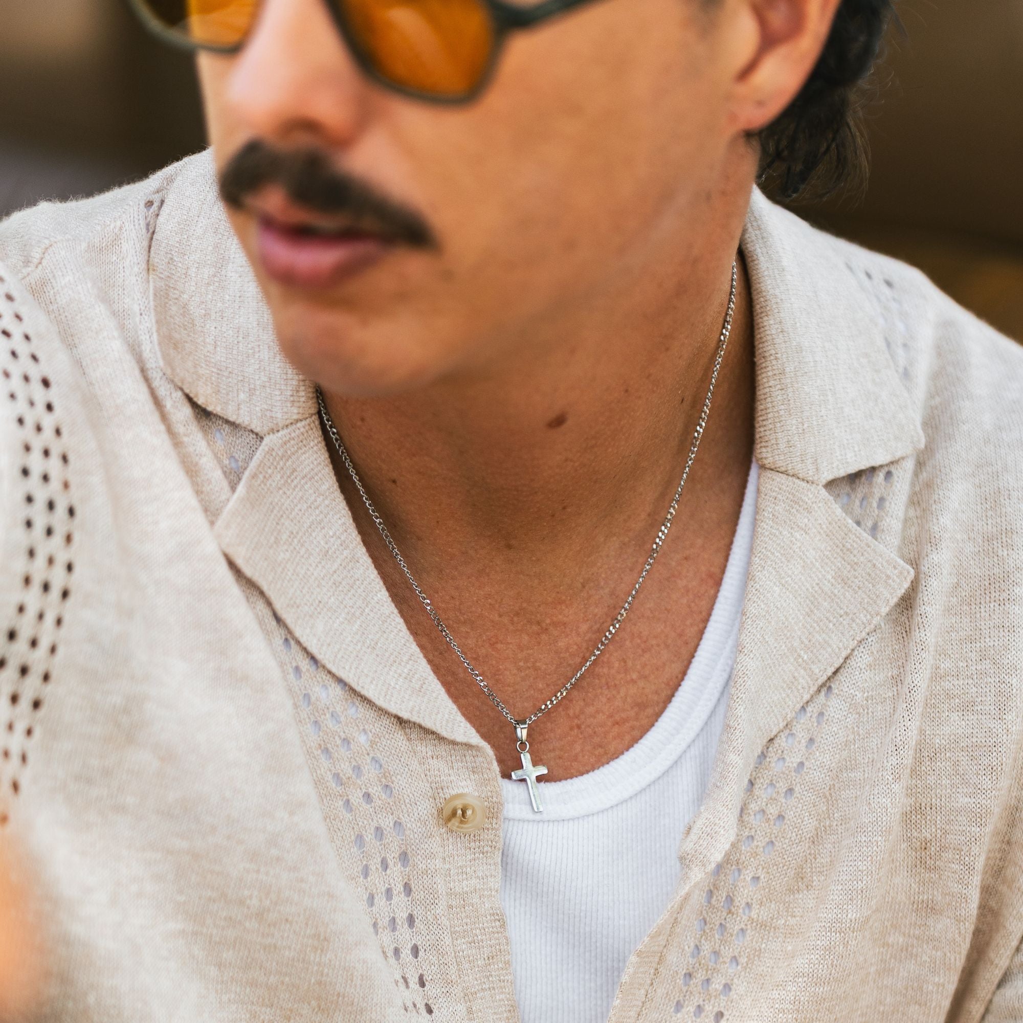A close-up lifestyle shot of a man wearing a silver cross pendant necklace over a white t-shirt and a beige linen button-up shirt.