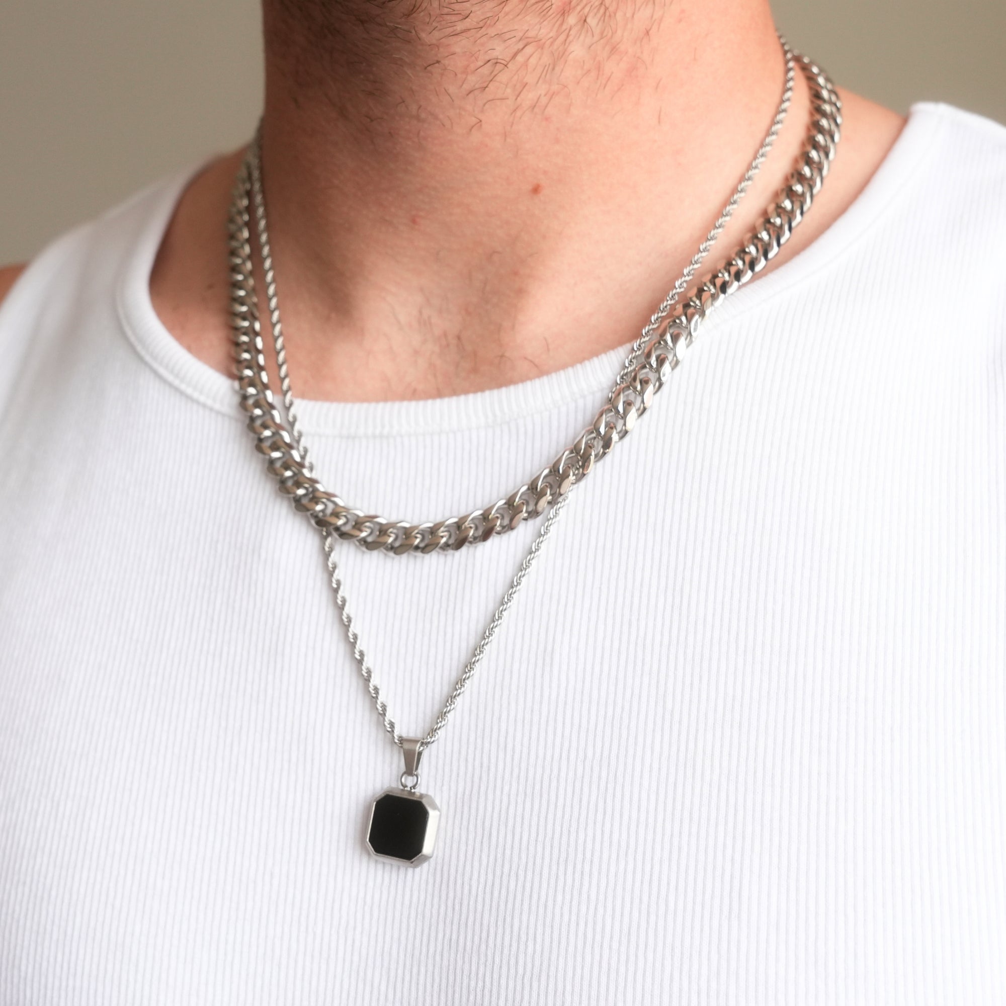 Person wearing two silver chain necklaces with a black pendant on a plain background