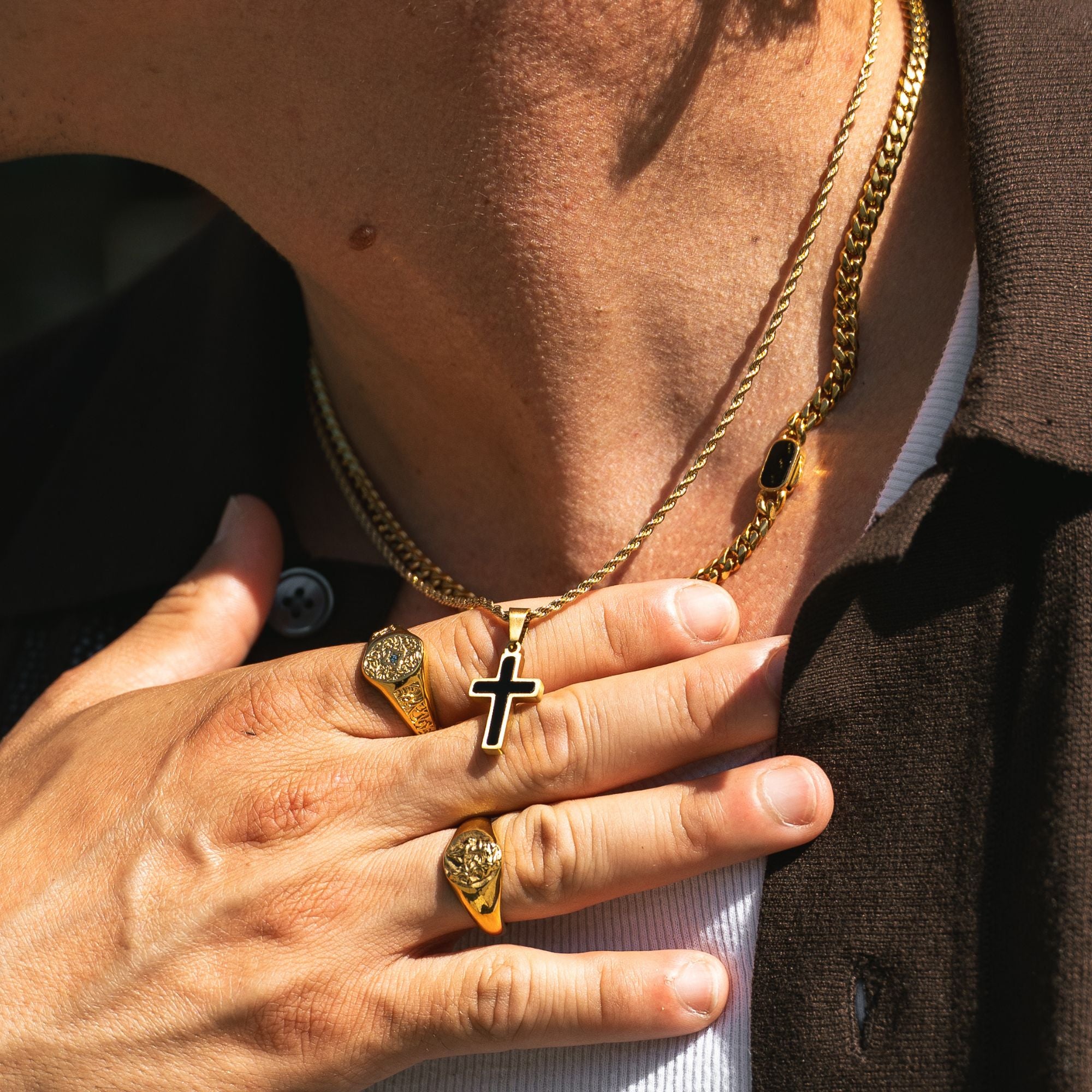 A close-up lifestyle shot of a person wearing gold jewelry, featuring a gold Cuban link chain necklace with a black enamel cross pendant, a gold rope chain necklace with a black rectangular stone, and gold signet rings on the fingers.
