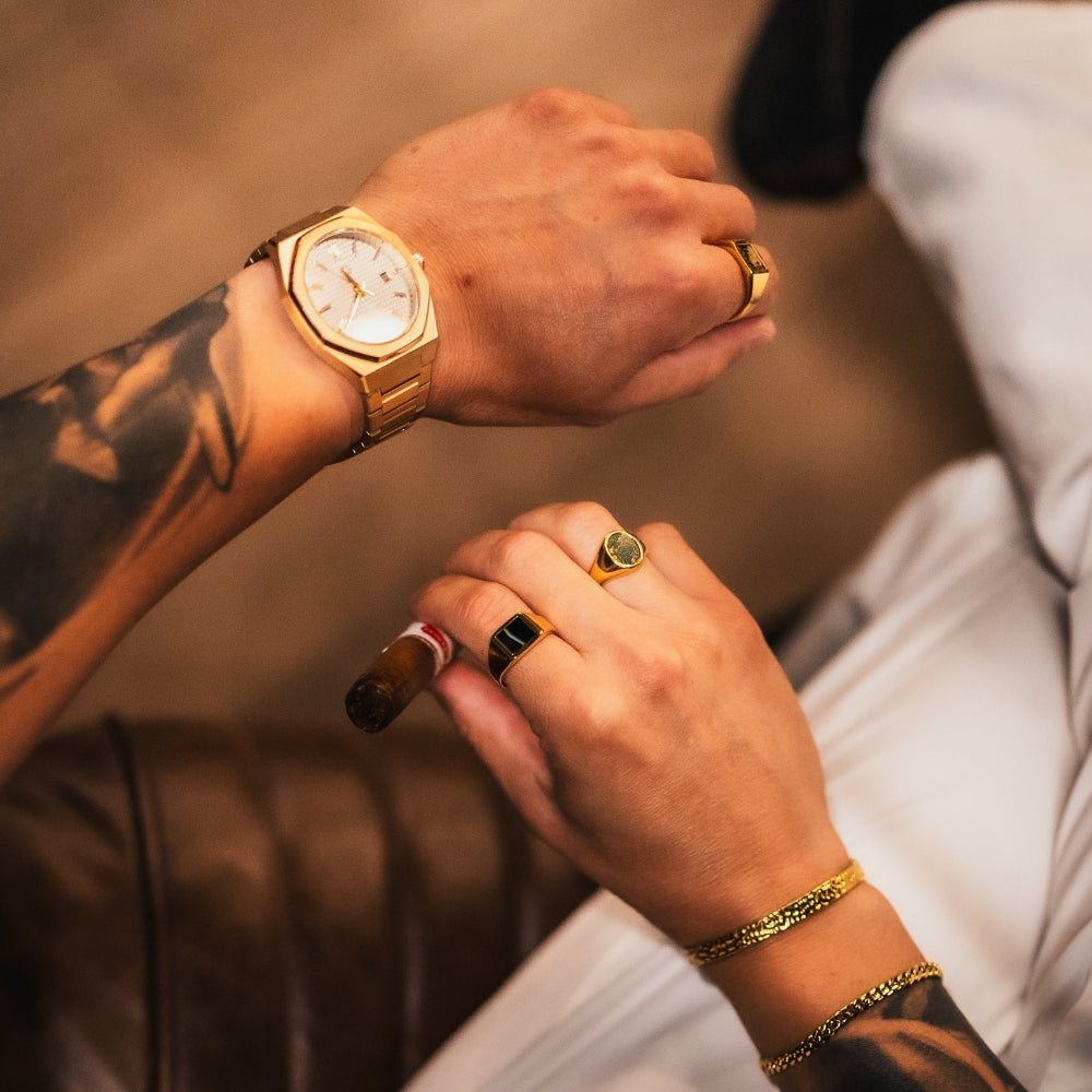 Close-up of a person's hands with tattoos, wearing a gold watch and rings, holding a cigar.