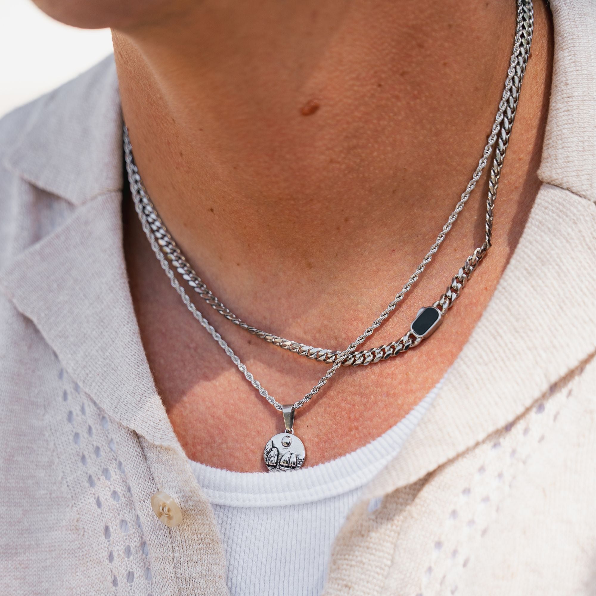 A close-up of a person's neck wearing a layered necklace set. The set includes a thick silver curb chain, a thinner silver chain with a small rectangular black stone accent, and a delicate silver chain with a round engraved pendant.