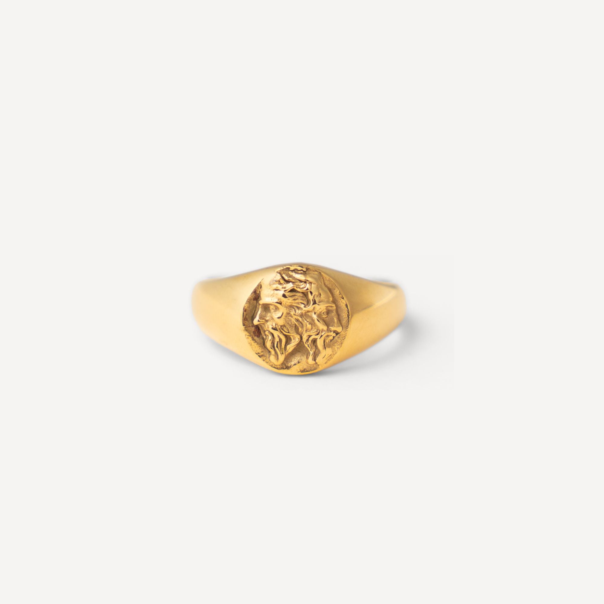 A gold signet ring featuring an embossed circular design with a stylized face, displayed on a clean, light gray background.