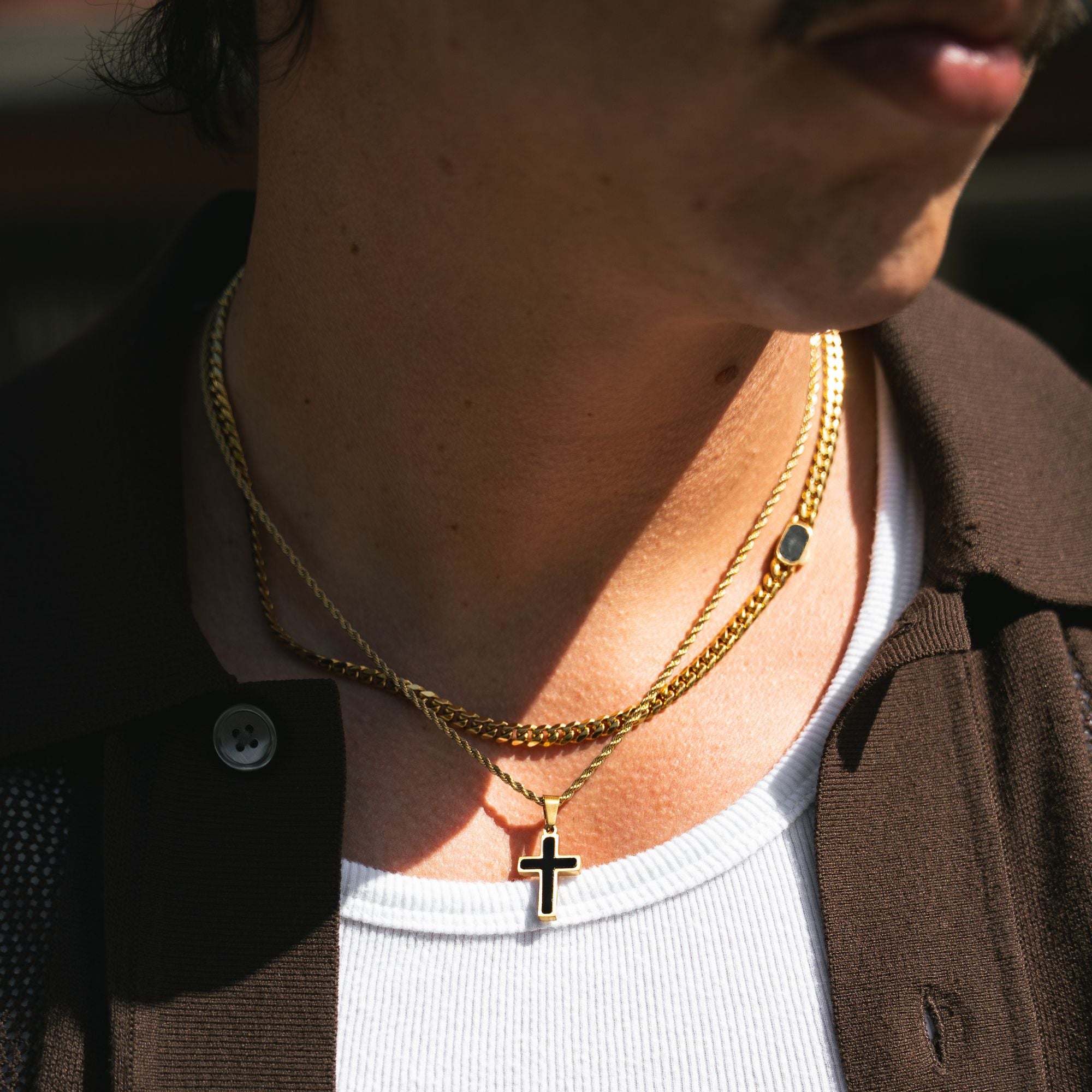 A close-up of a person's neck and chest wearing two layered gold necklaces, including a cross pendant, over a white t-shirt and brown jacket.