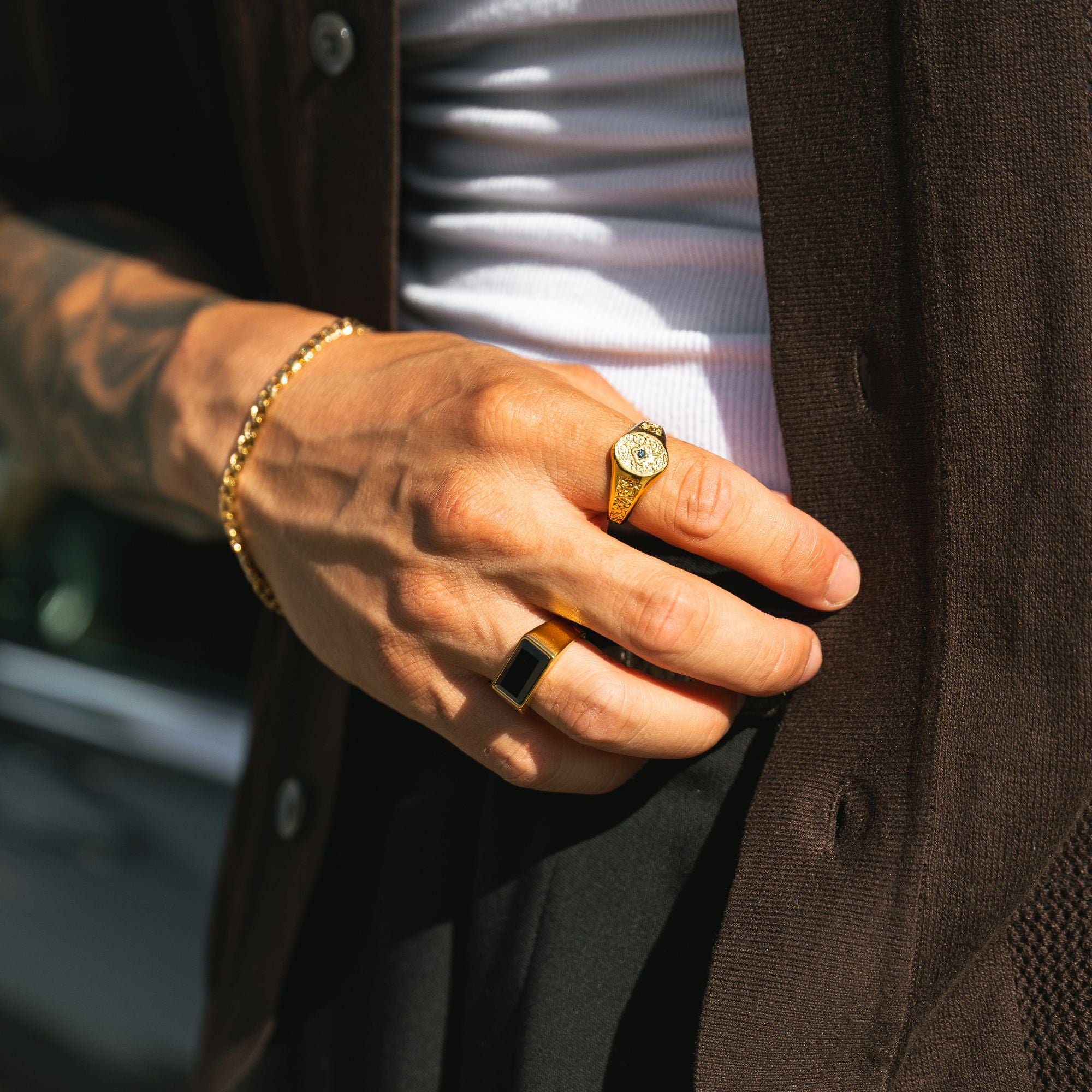 A close-up lifestyle shot of a man's hand wearing gold jewelry, including a gold signet ring, a gold band ring, and a gold chain bracelet, styled with a brown cardigan and white shirt.