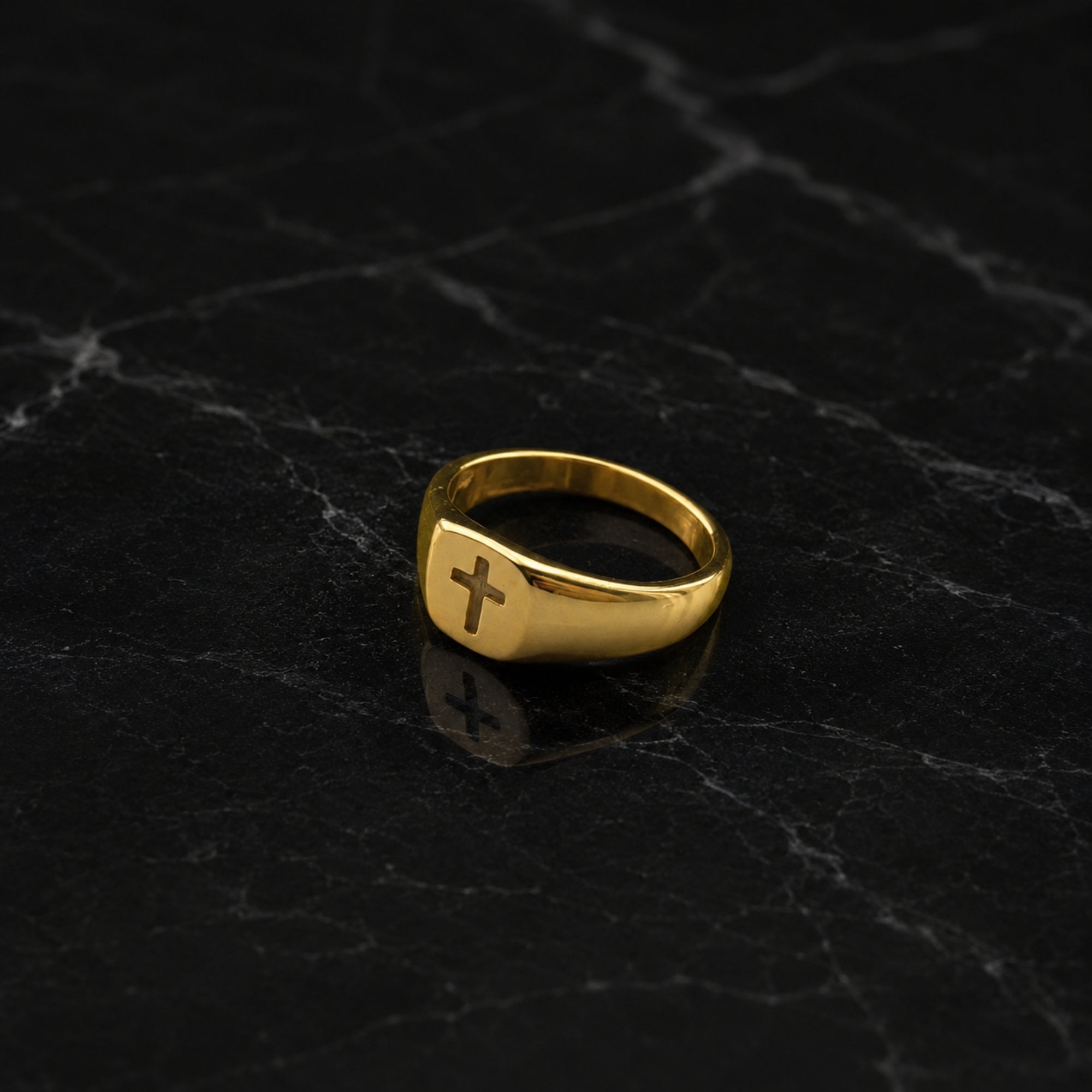 A polished gold signet ring with a raised cross emblem on the face, displayed on a dark black marble surface.
