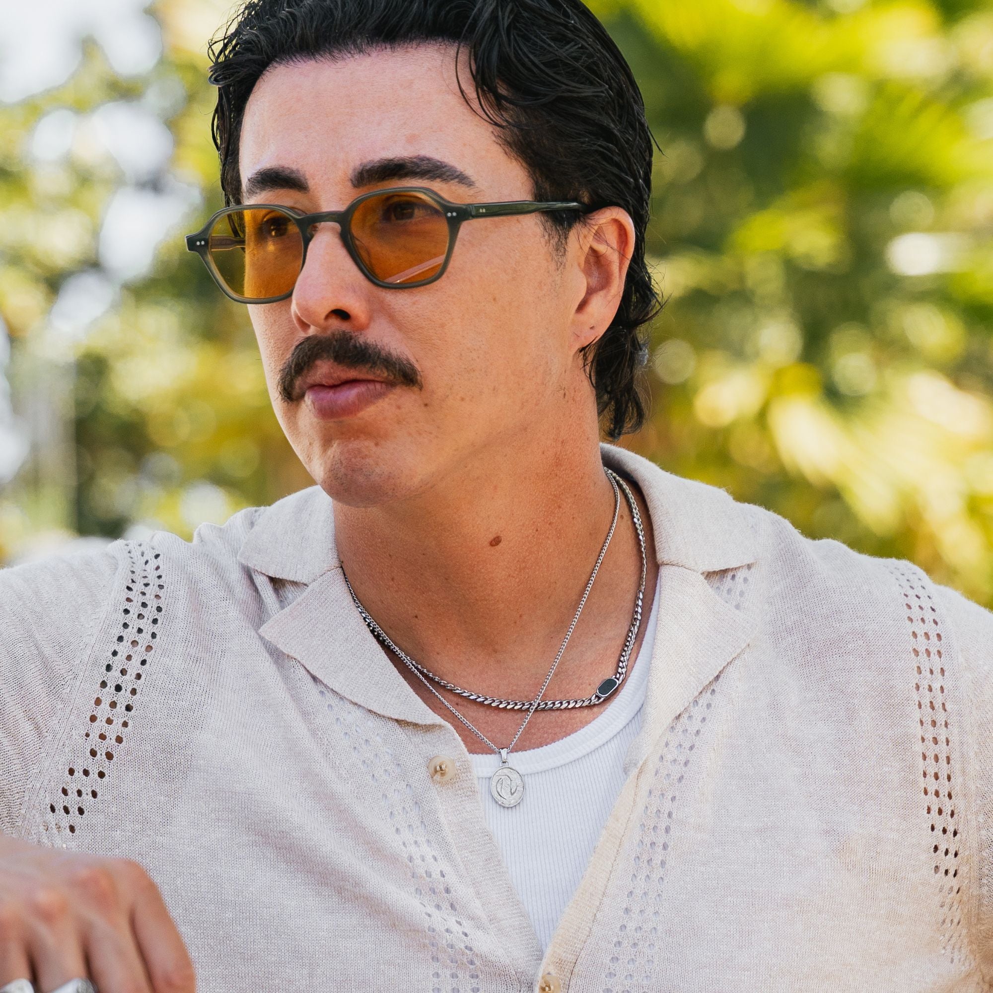 A close-up portrait of a man wearing distinctive geometric sunglasses with amber-tinted lenses and dark green frames, styled outdoors with a blurred green background.