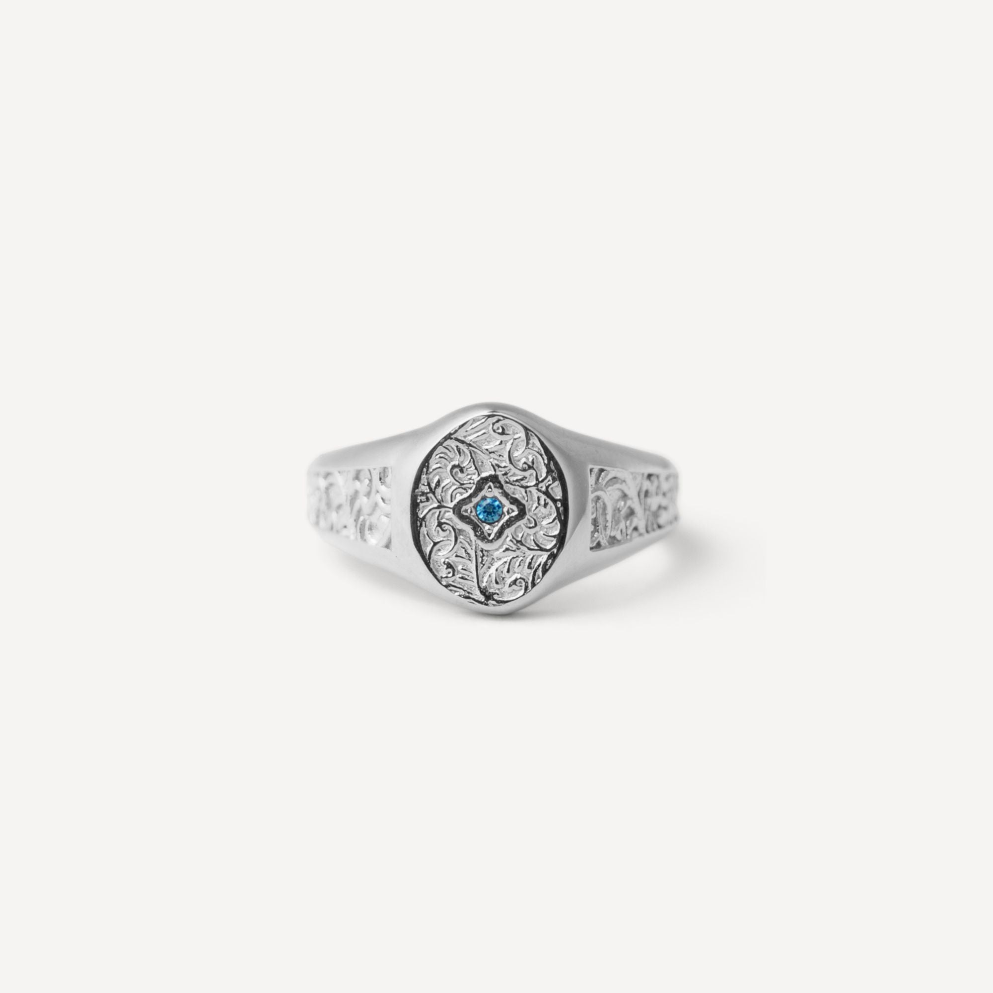 A silver signet ring with an ornate floral engraving on the band and a central oval face featuring a small blue star motif, set against a plain light gray background.