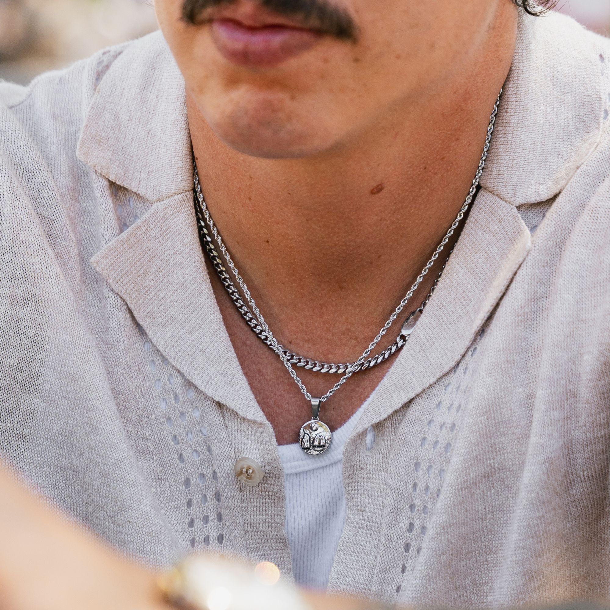 A close-up of a man wearing layered silver necklaces, including a curb chain and a rope chain with a small circular pendant, over a light beige linen shirt.