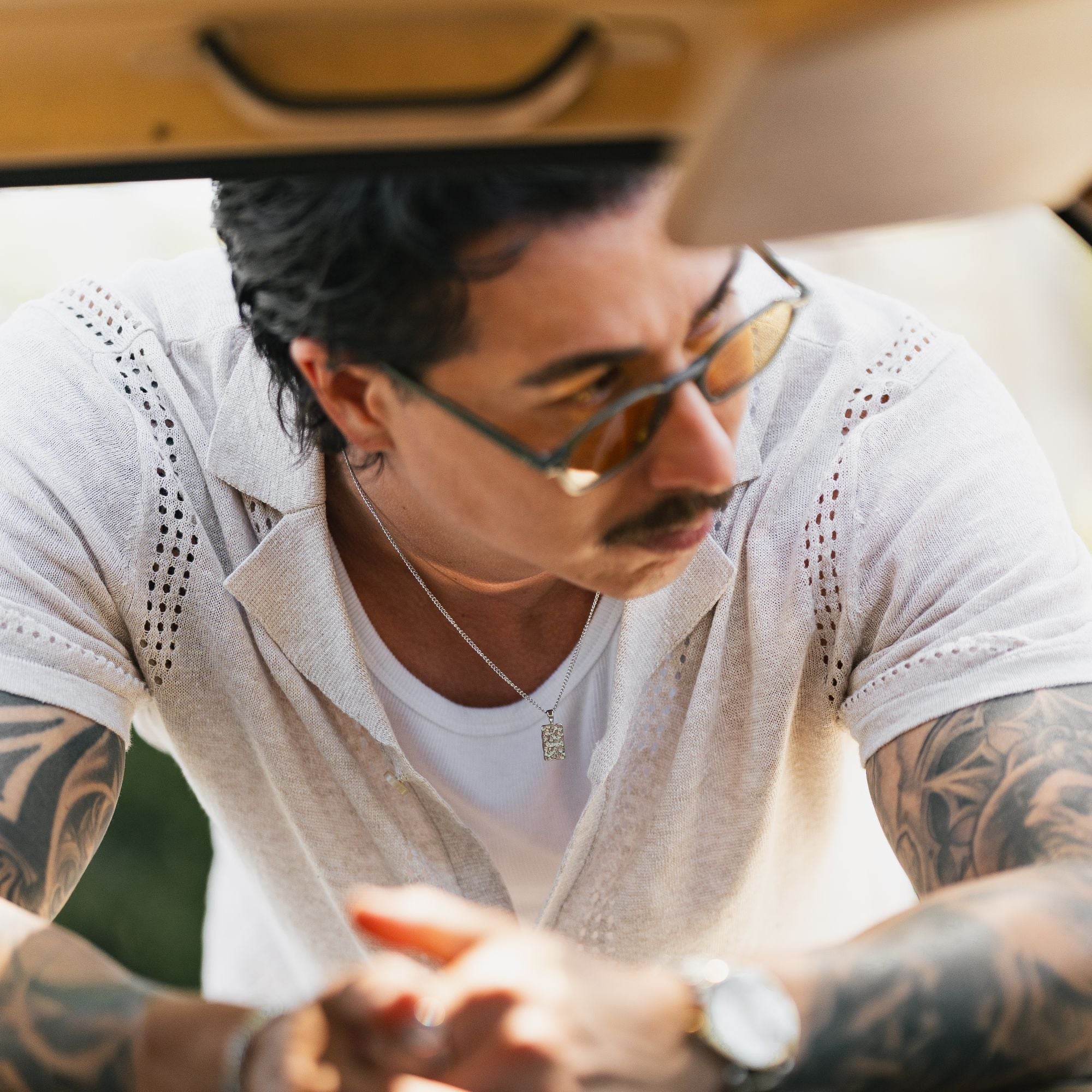 A close-up lifestyle shot of a man with tattoos wearing a white short-sleeve button-up shirt with eyelet detailing, a white t-shirt, and sunglasses, looking down in a sunny outdoor setting.