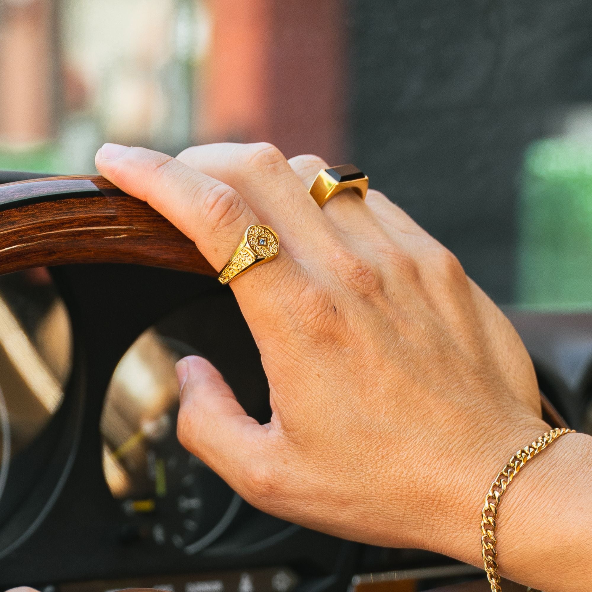 A close-up of a person's hand resting on a wooden car steering wheel, wearing a gold ring with a detailed floral design and a gold signet-style ring.
